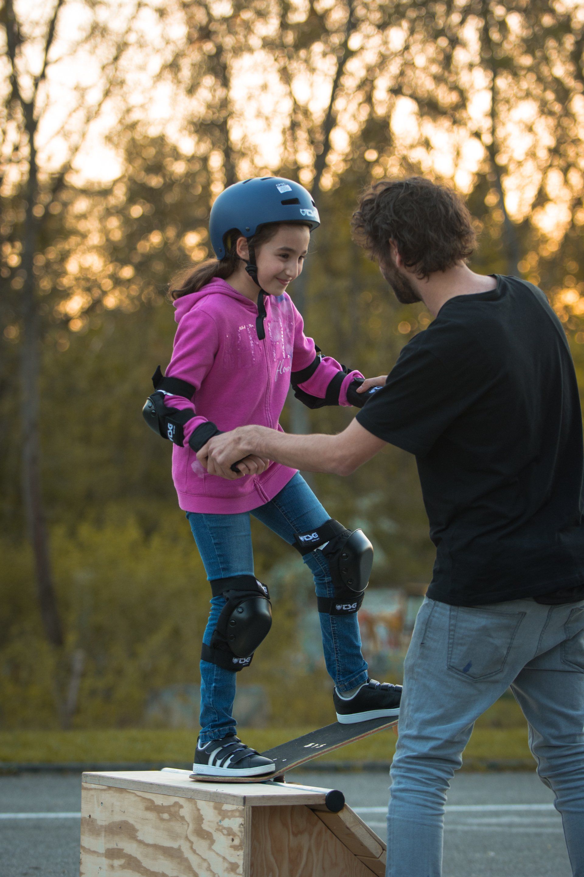 Mädchen in Schutzausrüstung lernt mit einem Lehrer auf einer Rampe Skateboardfahren. Sonnenuntergang im Hintergrund.