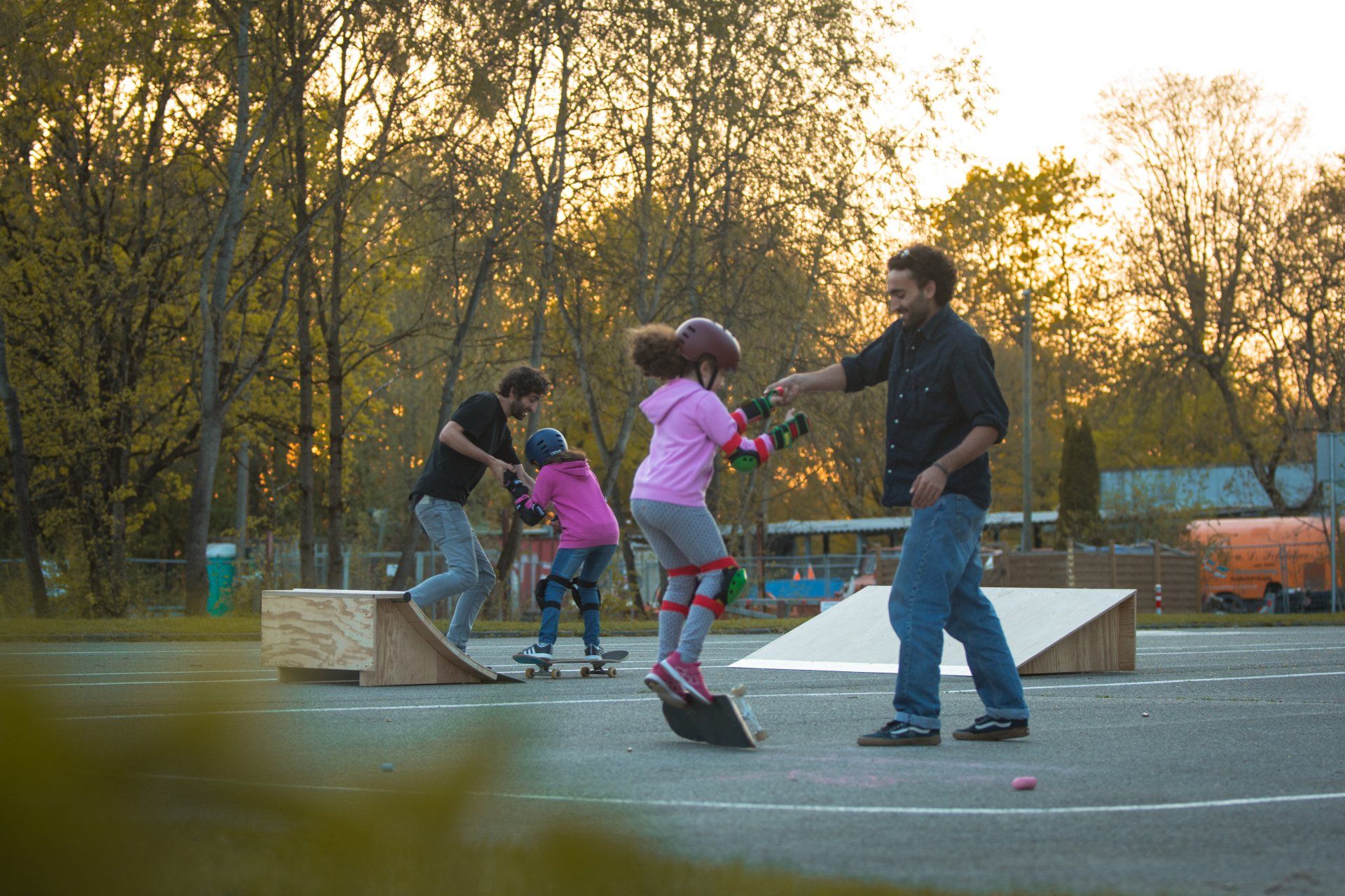 Erwachsene helfen Kindern beim Skateboarden in einem Skatepark; die untergehende Sonne wirft ein goldenes Licht.