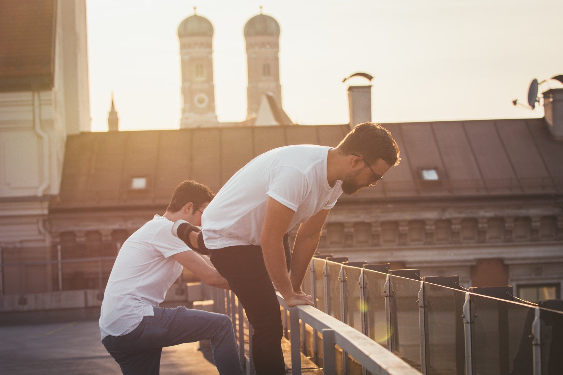 Zwei Männer auf einem Dach, einer hilft dem anderen beim Überklettern eines Geländers, im Hintergrund die untergehende Sonne.
