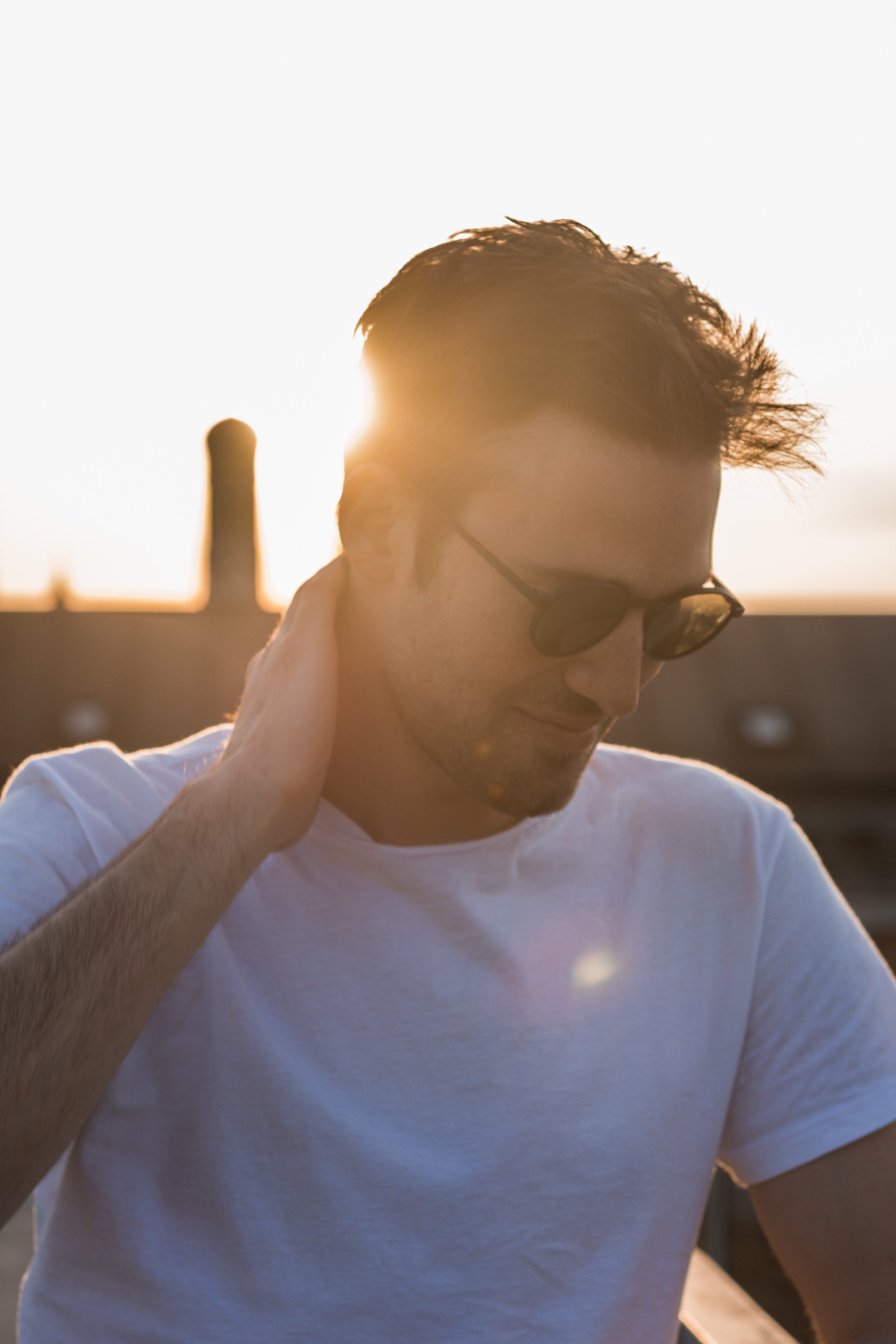 Mann mit Sonnenbrille berührt seinen Hals und steht im Sonnenlicht im Freien.