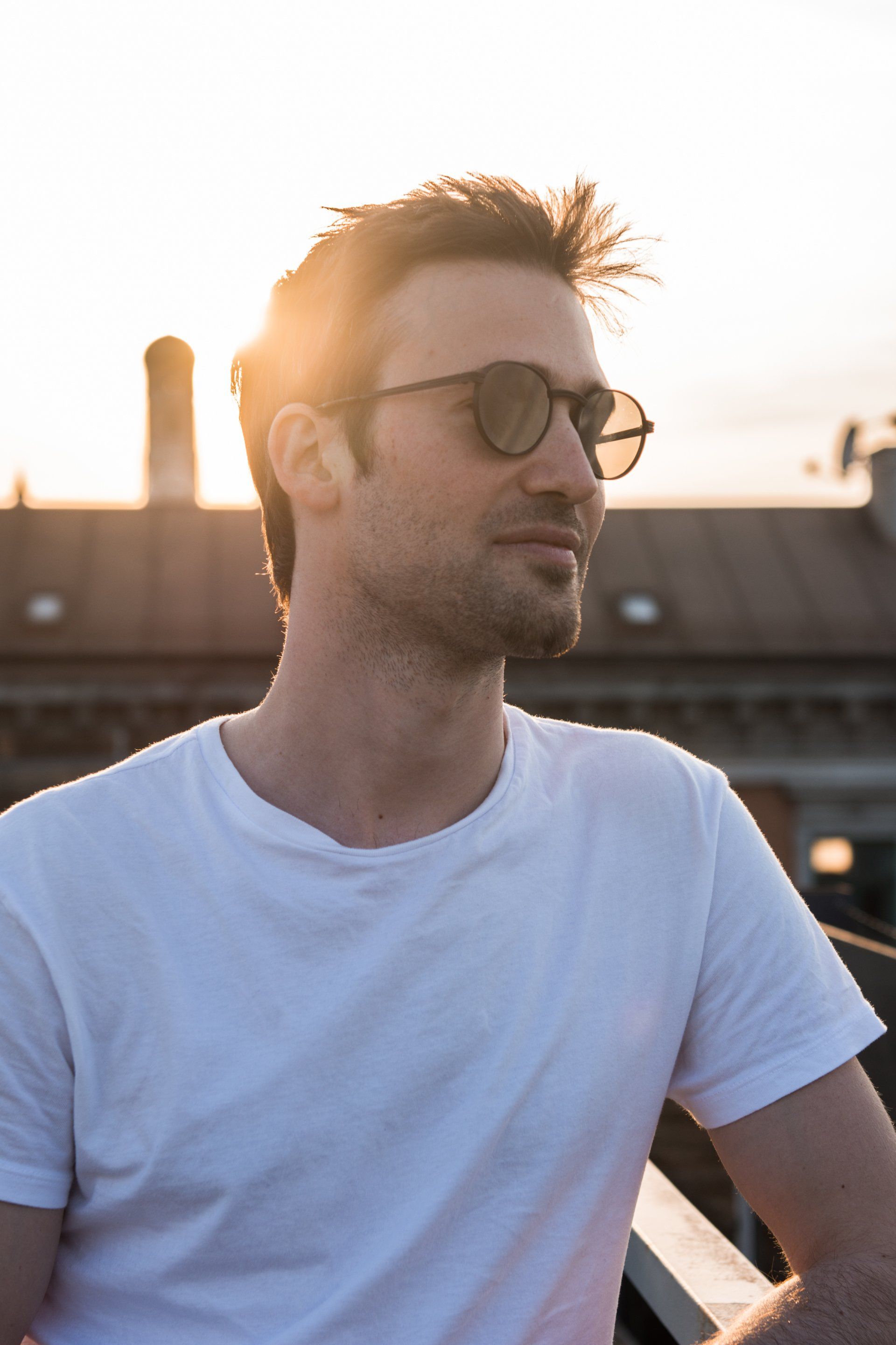 Mann mit Sonnenbrille auf einem Dach bei Sonnenuntergang, trägt ein weißes T-Shirt.
