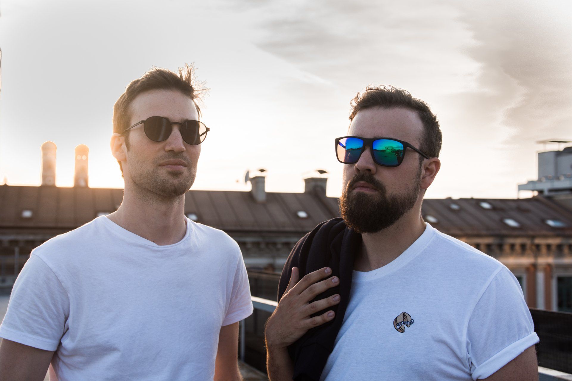 Zwei Männer mit Sonnenbrille stehen bei Sonnenuntergang auf einem Dach. 