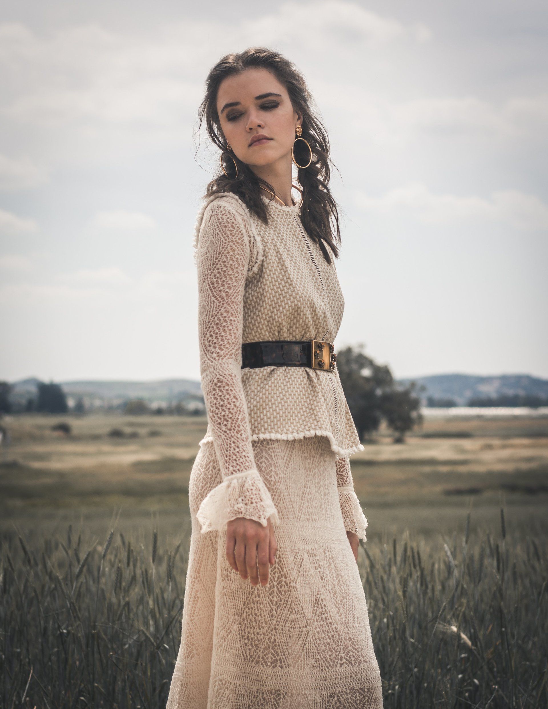 Frau in cremefarbenem Spitzenoutfit steht auf einem Feld, trägt einen schwarzen Gürtel, nachdenklicher Gesichtsausdruck.