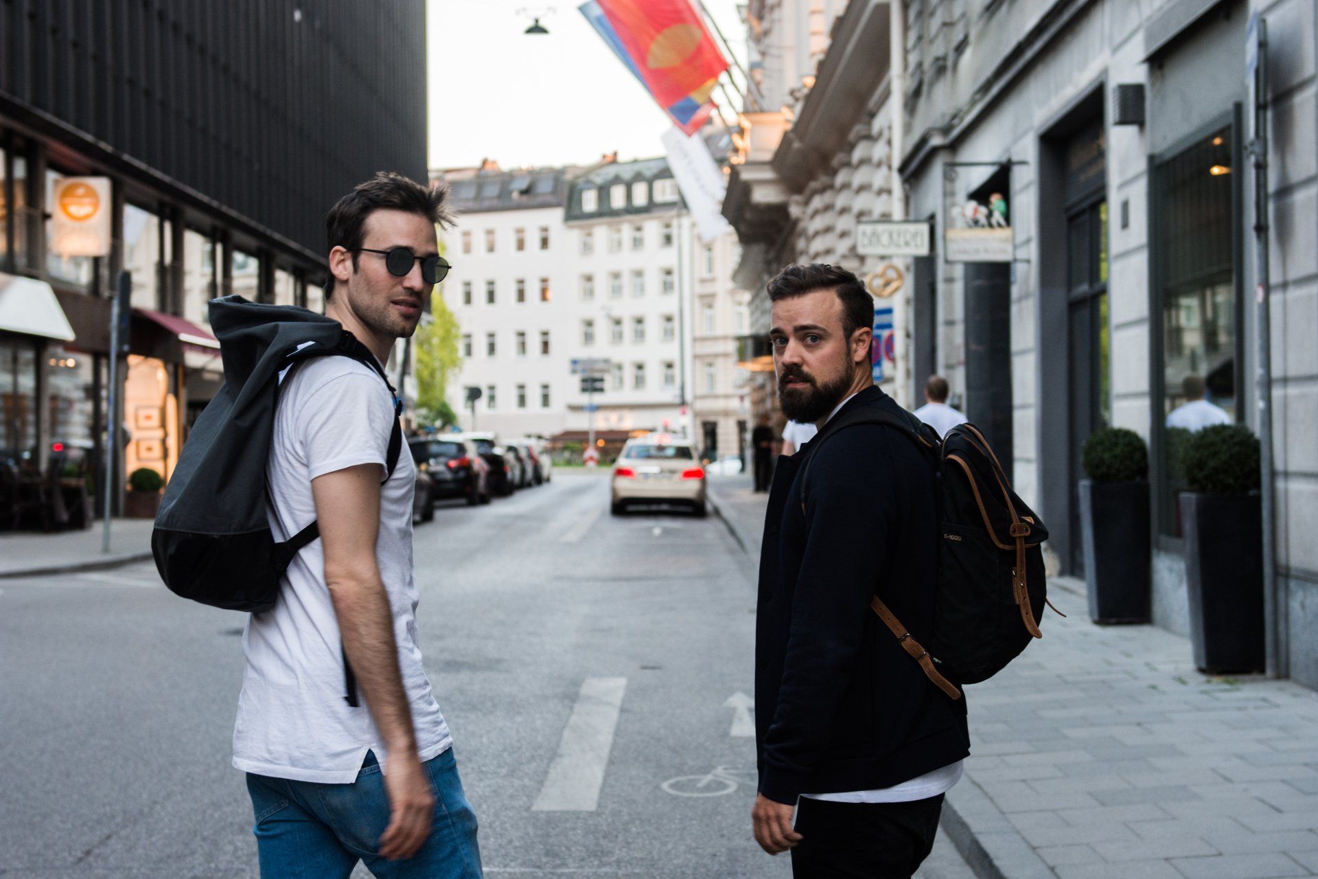 Zwei Männer gehen auf einer Stadtstraße und blicken in die Kamera. Sonnenlicht, Gebäude und Autos im Hintergrund.