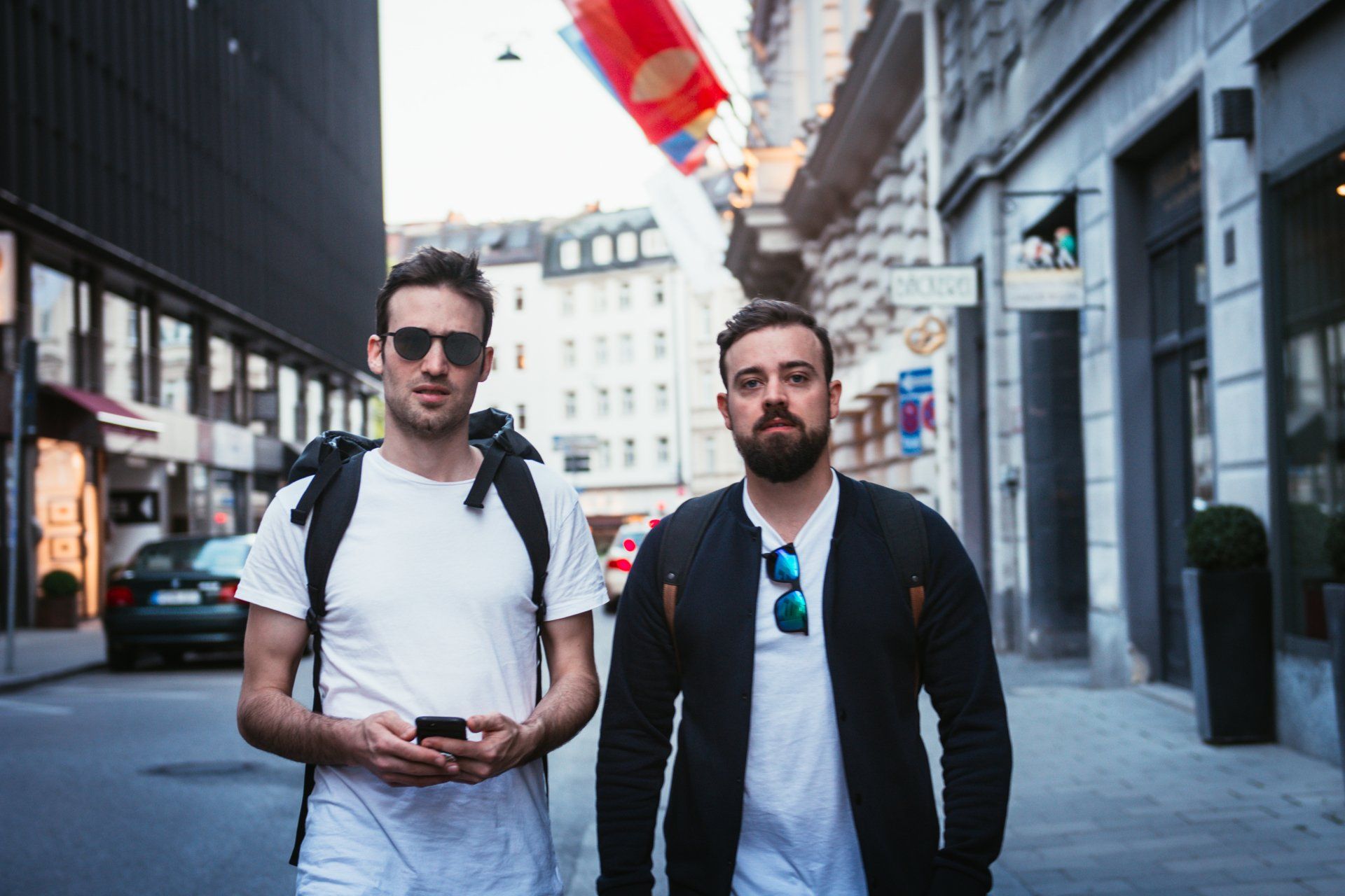 Zwei Männer gehen eine Stadtstraße entlang, einer mit Rucksack und Sonnenbrille, der andere mit Jacke und Sonnenbrille.