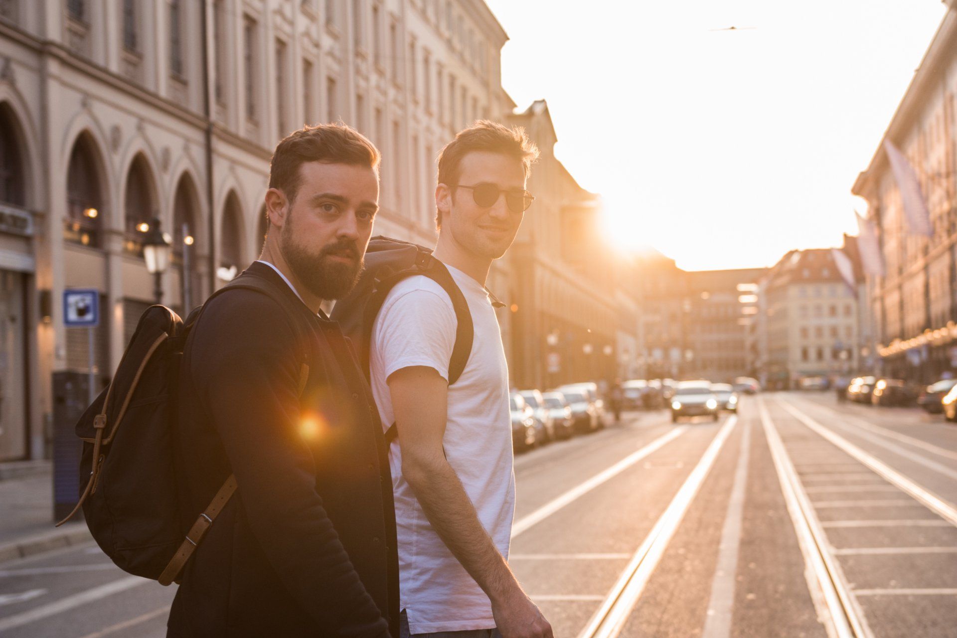Zwei Männer überqueren bei Sonnenuntergang eine Straße; einer mit Bart, Jacke und Rucksack.