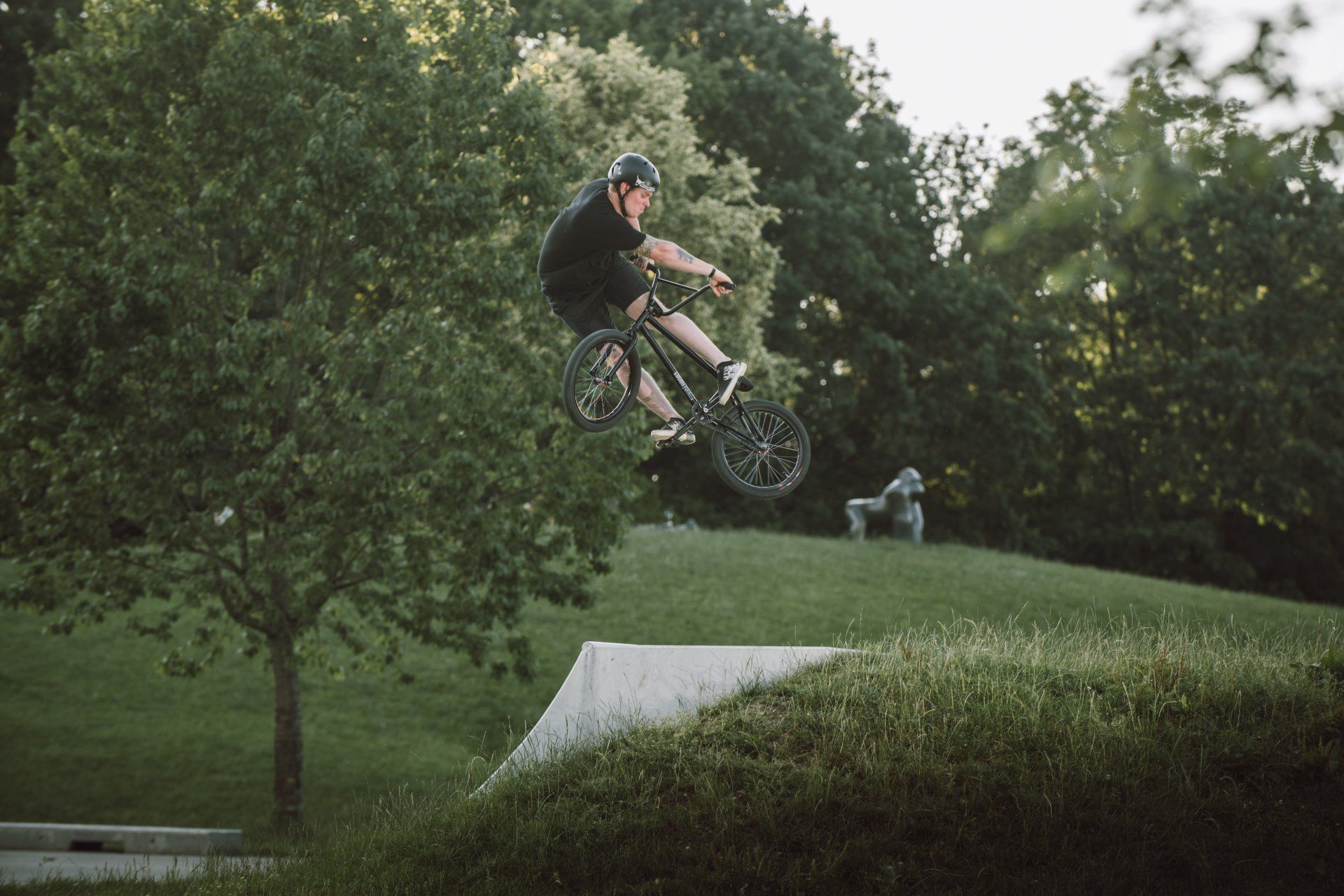 BMX-Fahrer in der Luft über einer Rampe in einem Park, umgeben von grünen Bäumen und Gras.