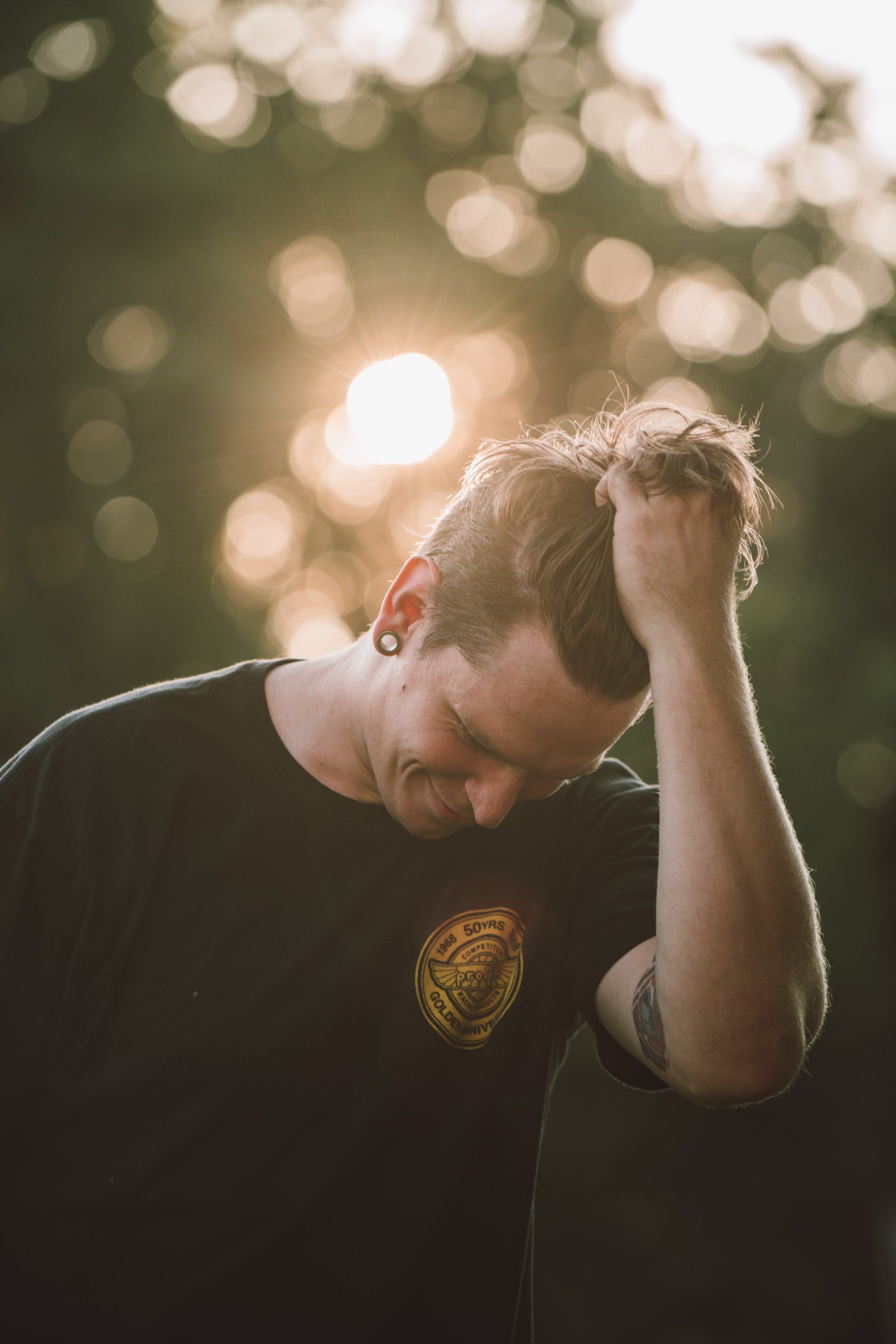 Mann im schwarzen Hemd, Hand im Haar, Blick nach unten, goldenes Sonnenlicht hinter ihm.