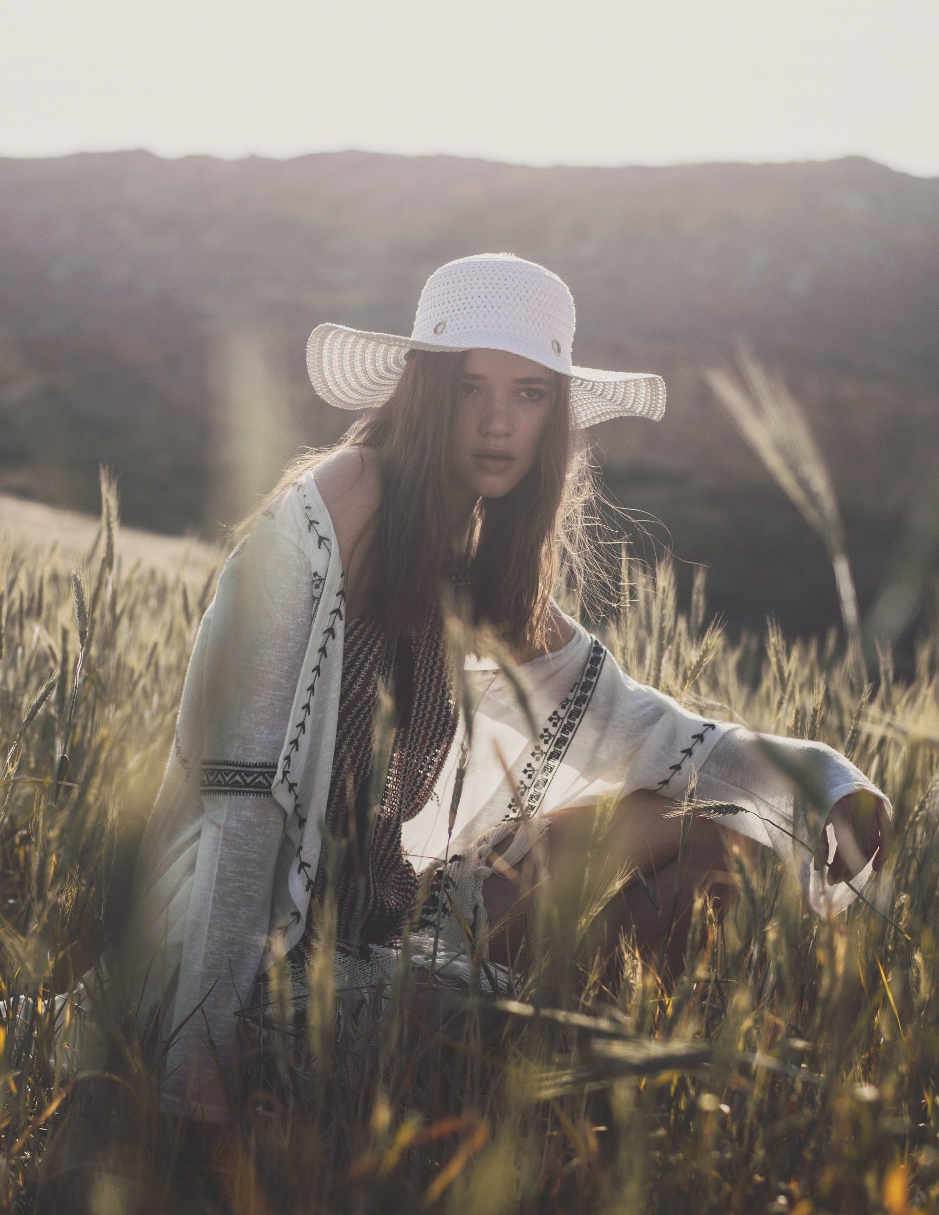Frau mit weißem Hut und Schal kniet in einem Weizenfeld und blickt mit leichtem Stirnrunzeln nach vorne.