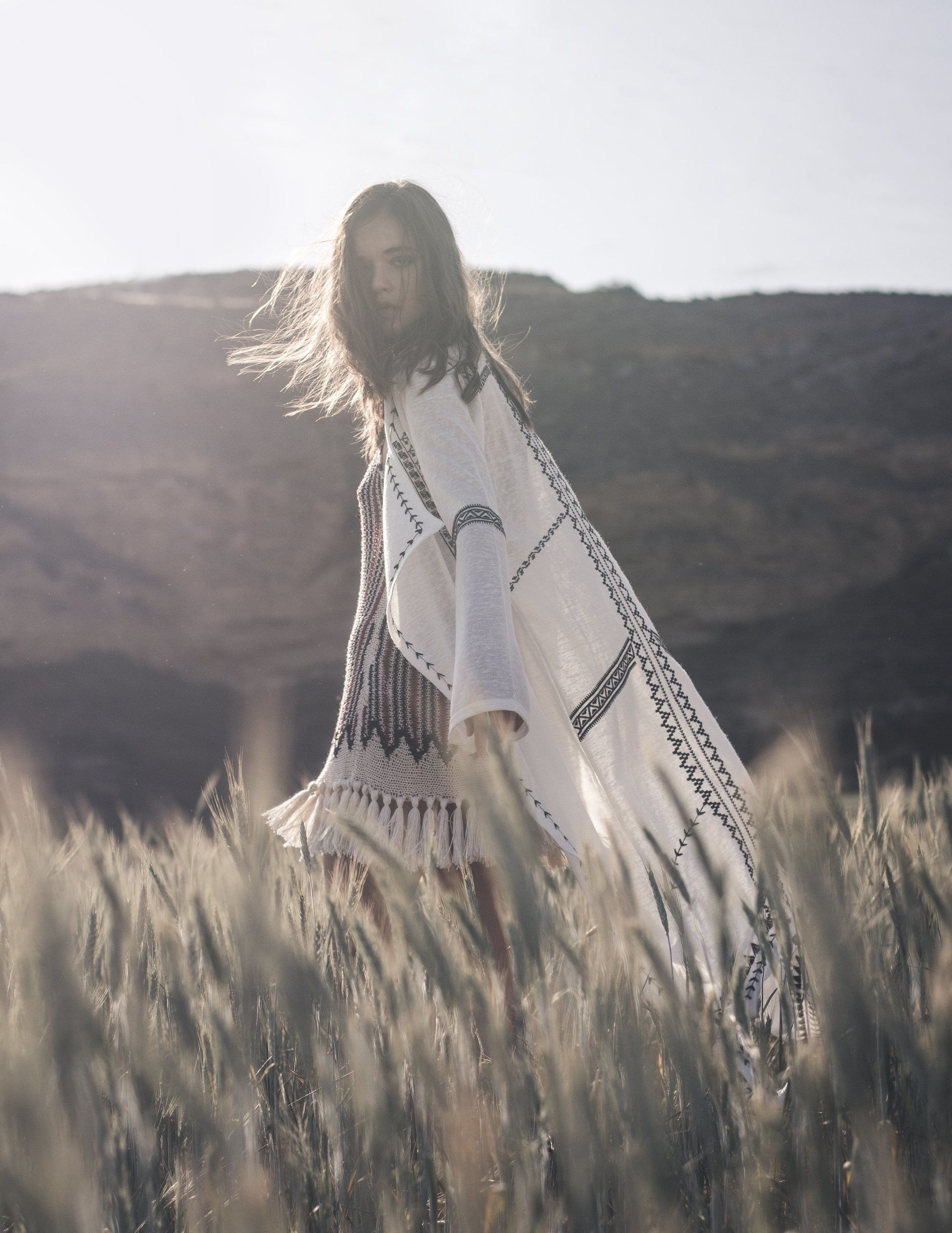 Frau im weißen bestickten Kimono geht durch ein Weizenfeld, Sonnenlicht, Berge im Hintergrund.