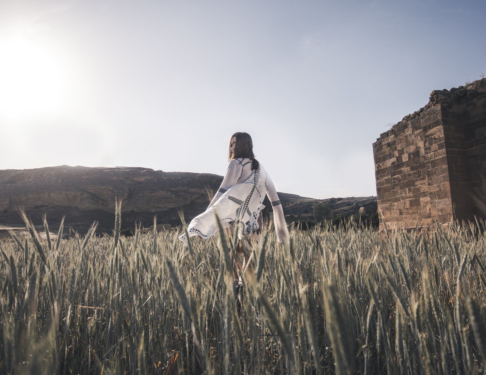 Frau im weißen Kleid geht im Sonnenlicht auf einem Feld spazieren, im Hintergrund eine alte Steinstruktur.