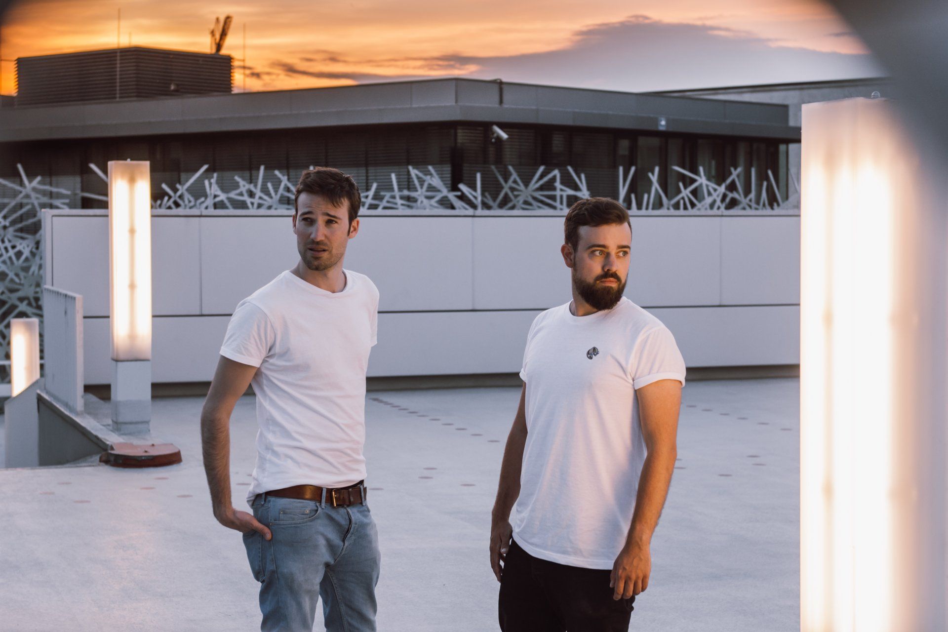 Zwei Männer in weißen T-Shirts stehen bei Sonnenuntergang auf einem Dach in der Nähe von vertikalen Lichtern.