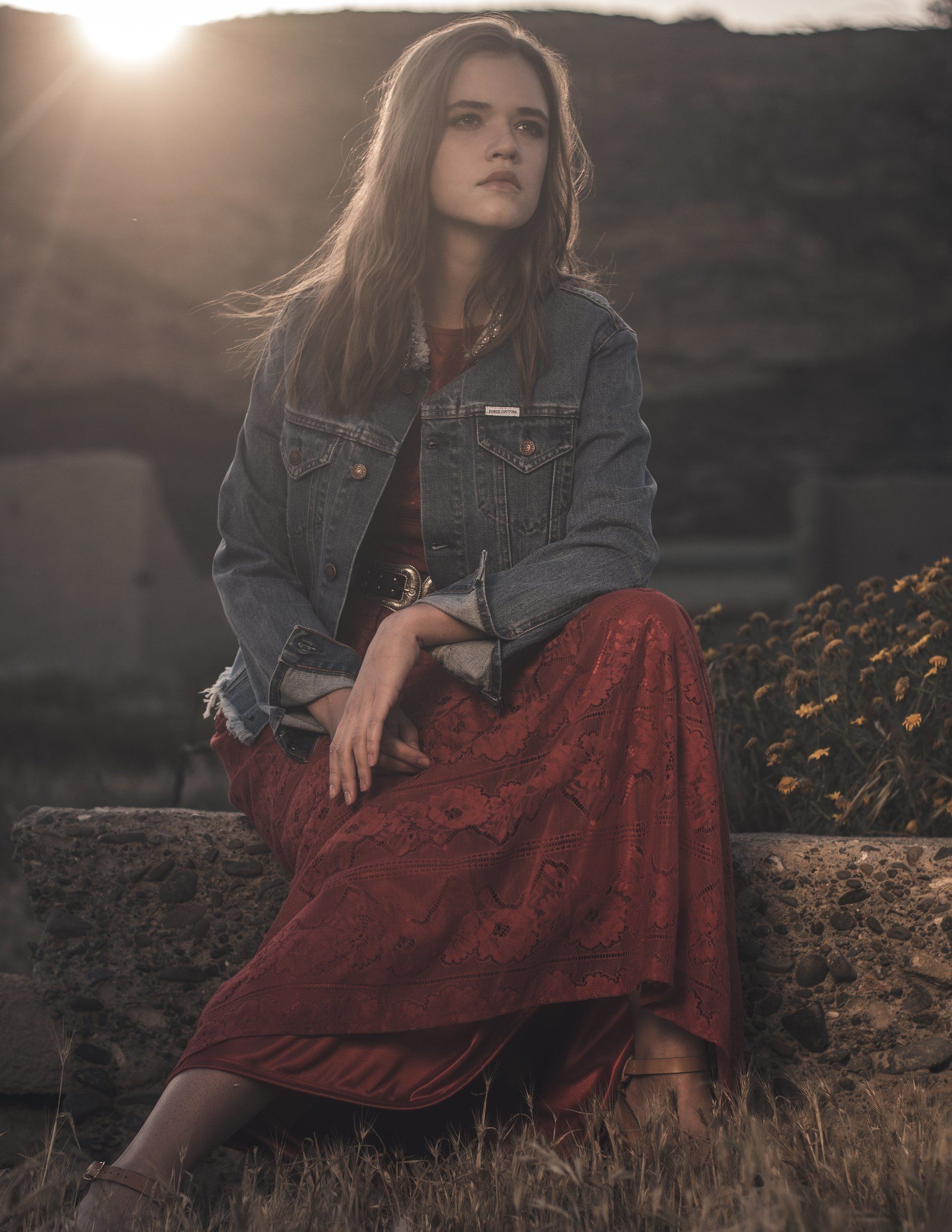 Frau im roten Kleid und Jeansjacke sitzt im Freien und sieht nachdenklich aus. Goldenes Sonnenlicht.