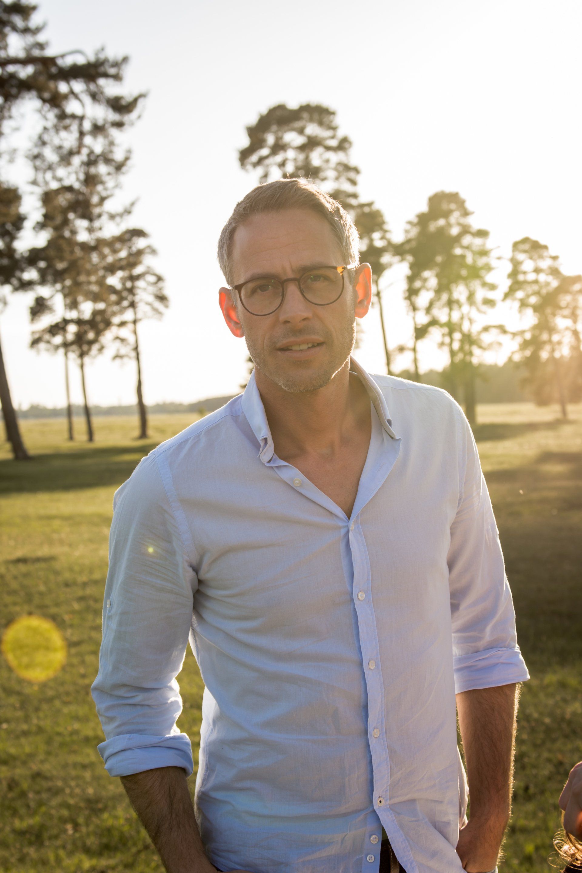 Mann mit Brille und hellblauem Hemd steht auf einem Feld mit Bäumen und Sonnenlicht im Hintergrund.