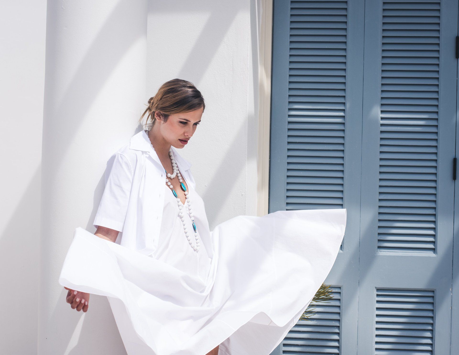 Frau in weißem Kleid und Halskette steht an einem sonnigen Tag neben blauen Fensterläden.
