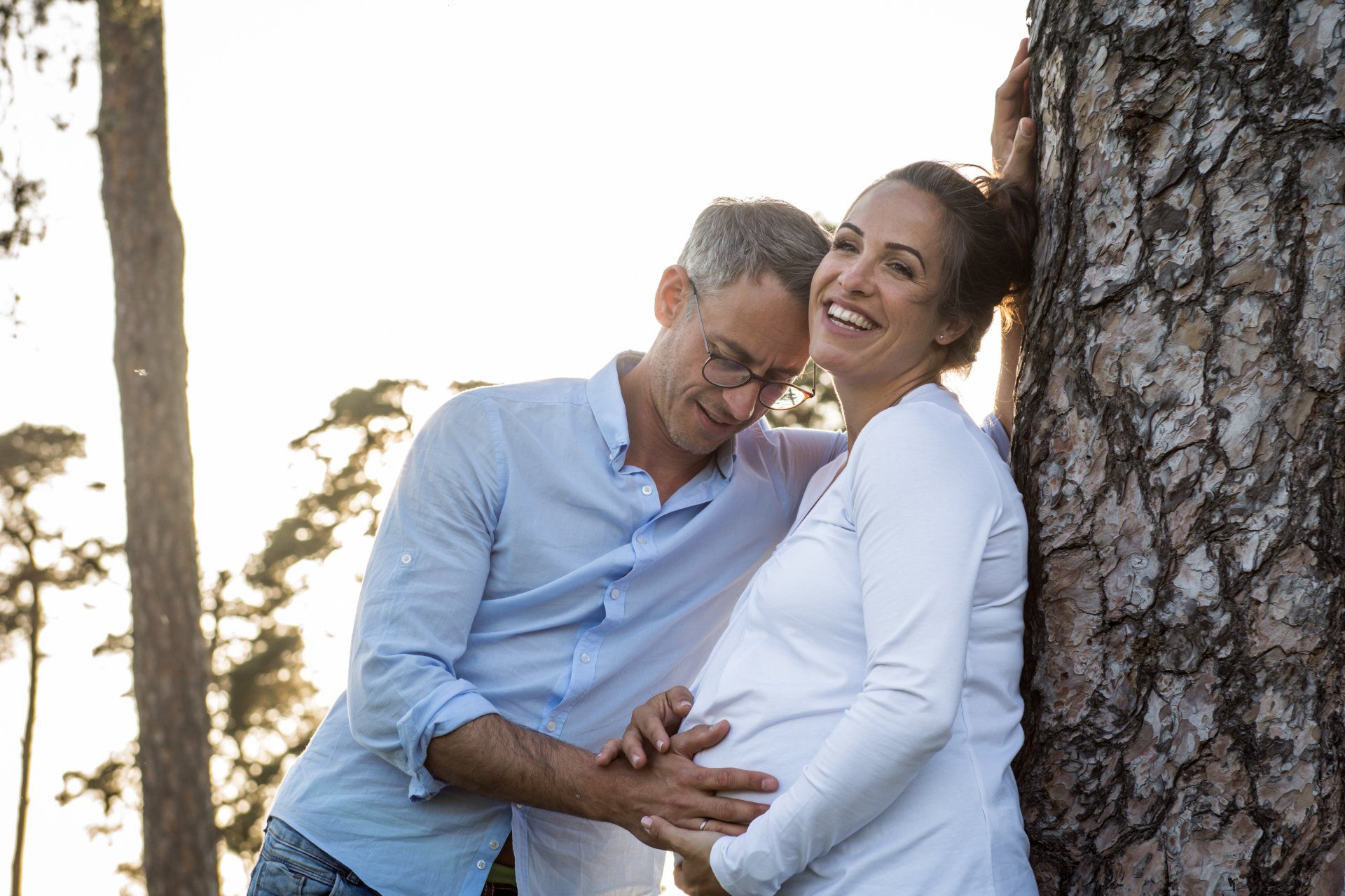 Schwangere Frau und Mann lächeln und berühren ihren Bauch in der Nähe eines Baumes. Sonnige Umgebung im Freien.