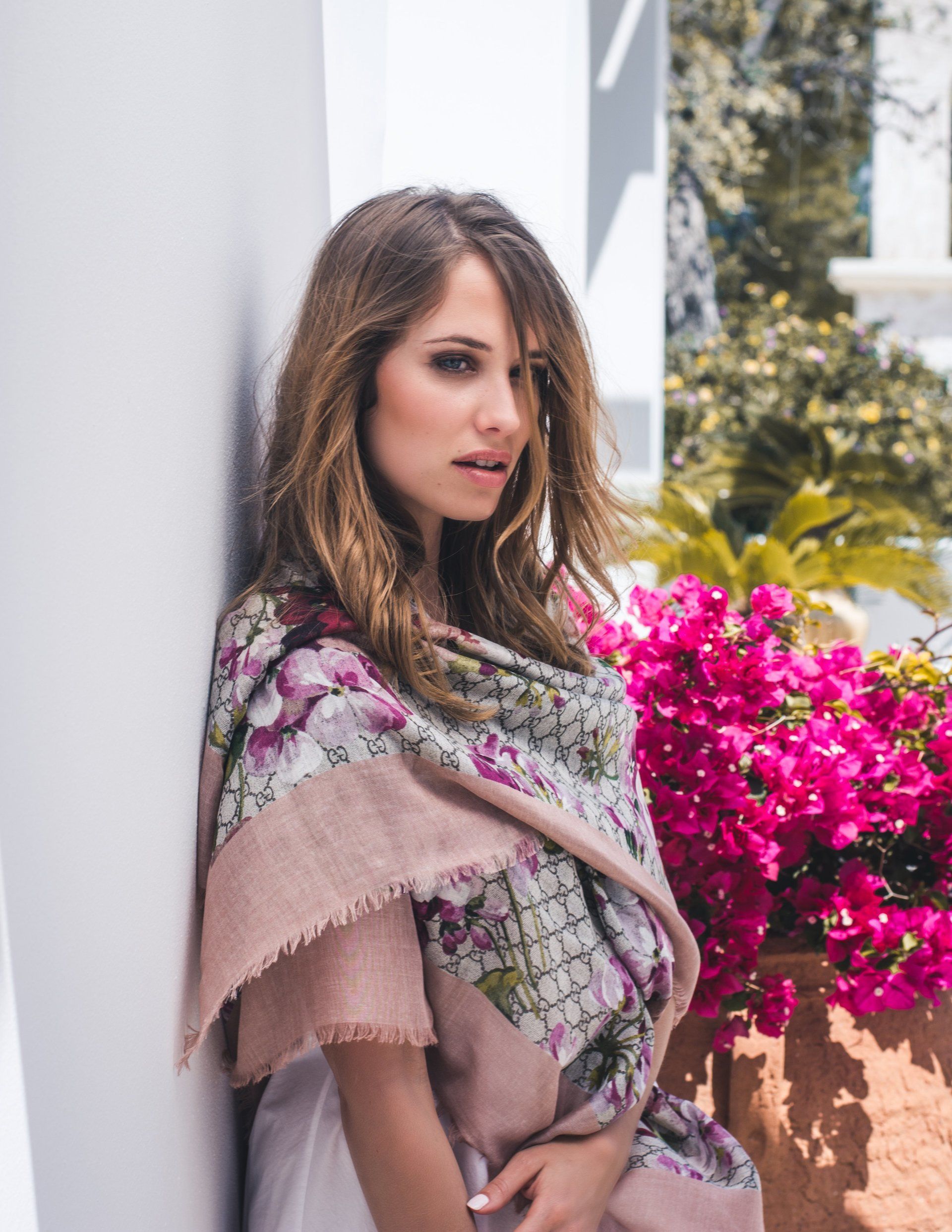 Frau lehnt an einer weißen Wand, trägt einen geblümten Schal, im Hintergrund sind leuchtend rosa Blumen zu sehen.