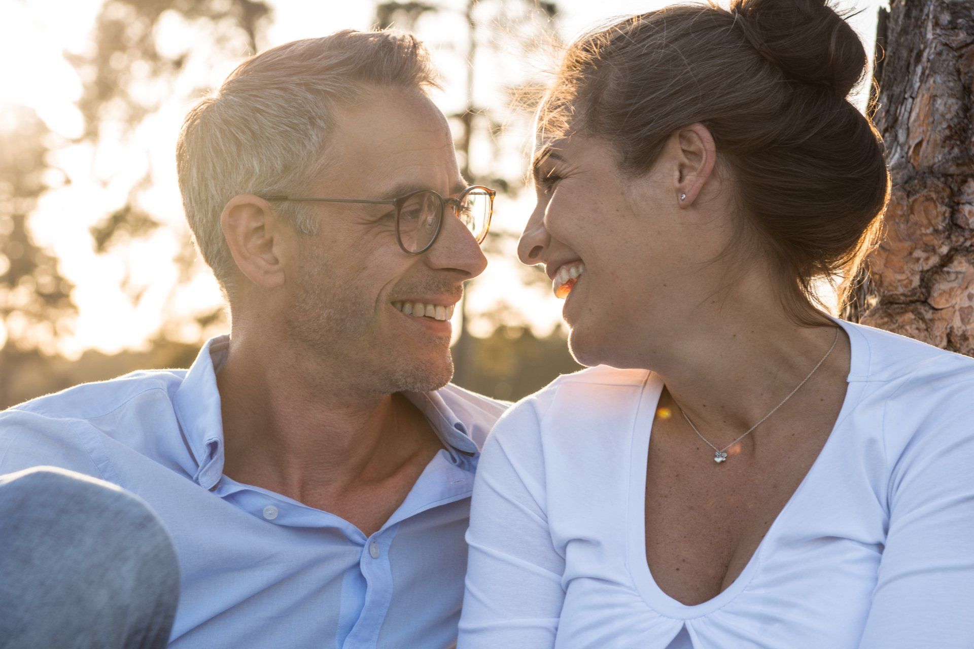 Lächelndes Paar, das sich anschaut; Frau mit hochgestecktem Haar, Mann mit Brille, Sonnenlicht im Hintergrund.