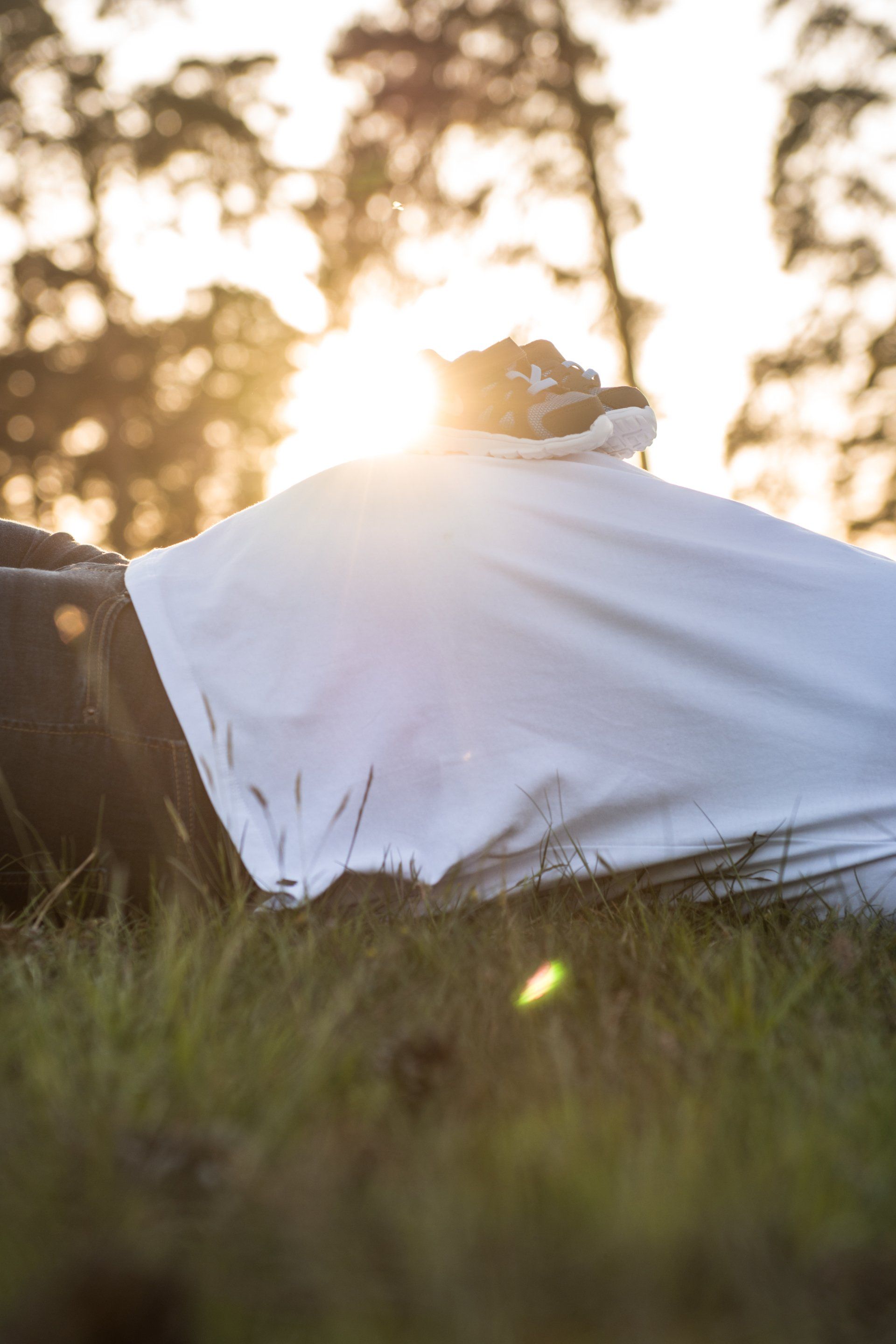 Person liegt im Gras, bedeckt mit einem weißen Laken. Helles Sonnenlicht fällt durch die Bäume im Hintergrund.