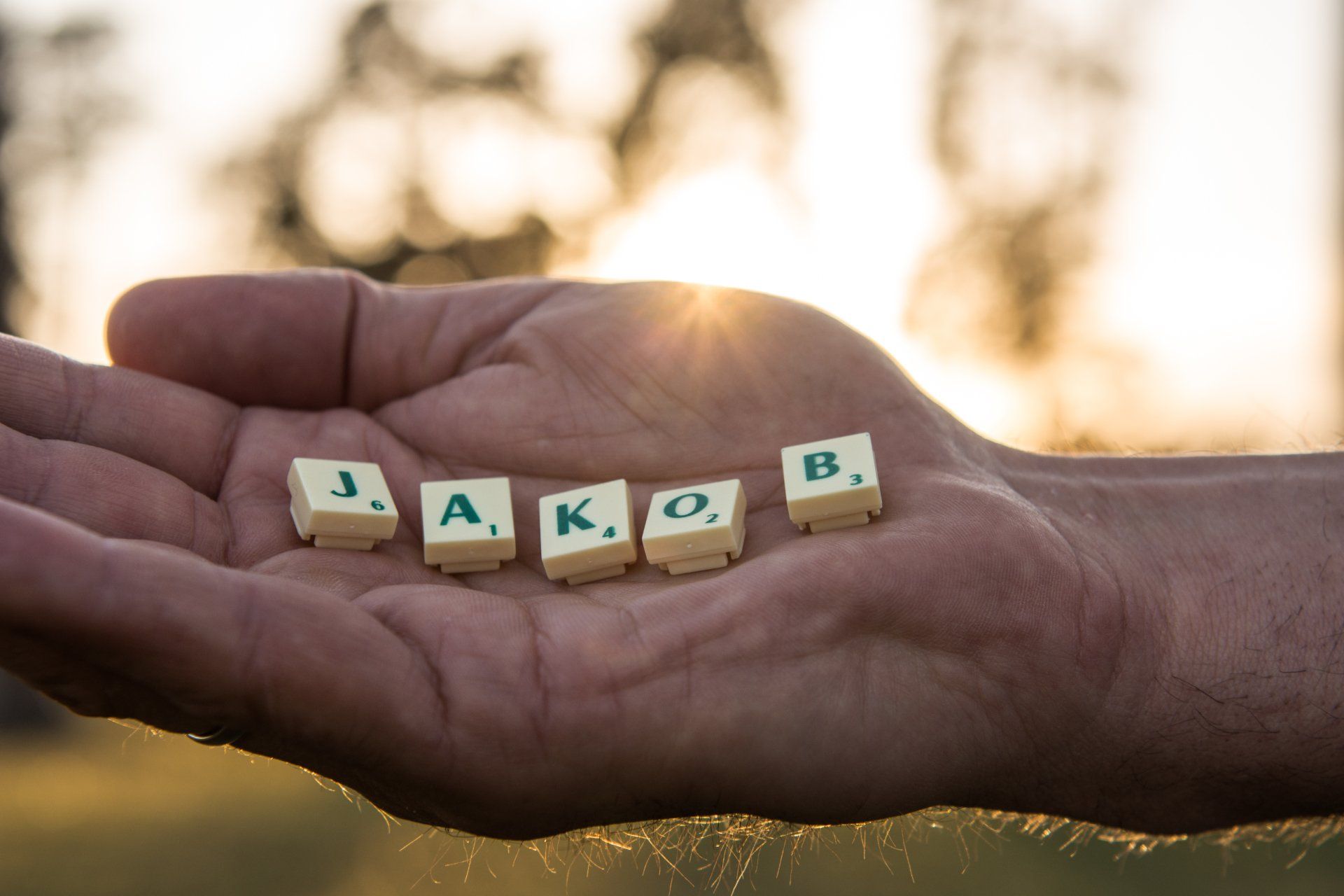 Hand hält Scrabble-Steine mit der Aufschrift „JAKOB“, Sonnenlicht im Hintergrund.