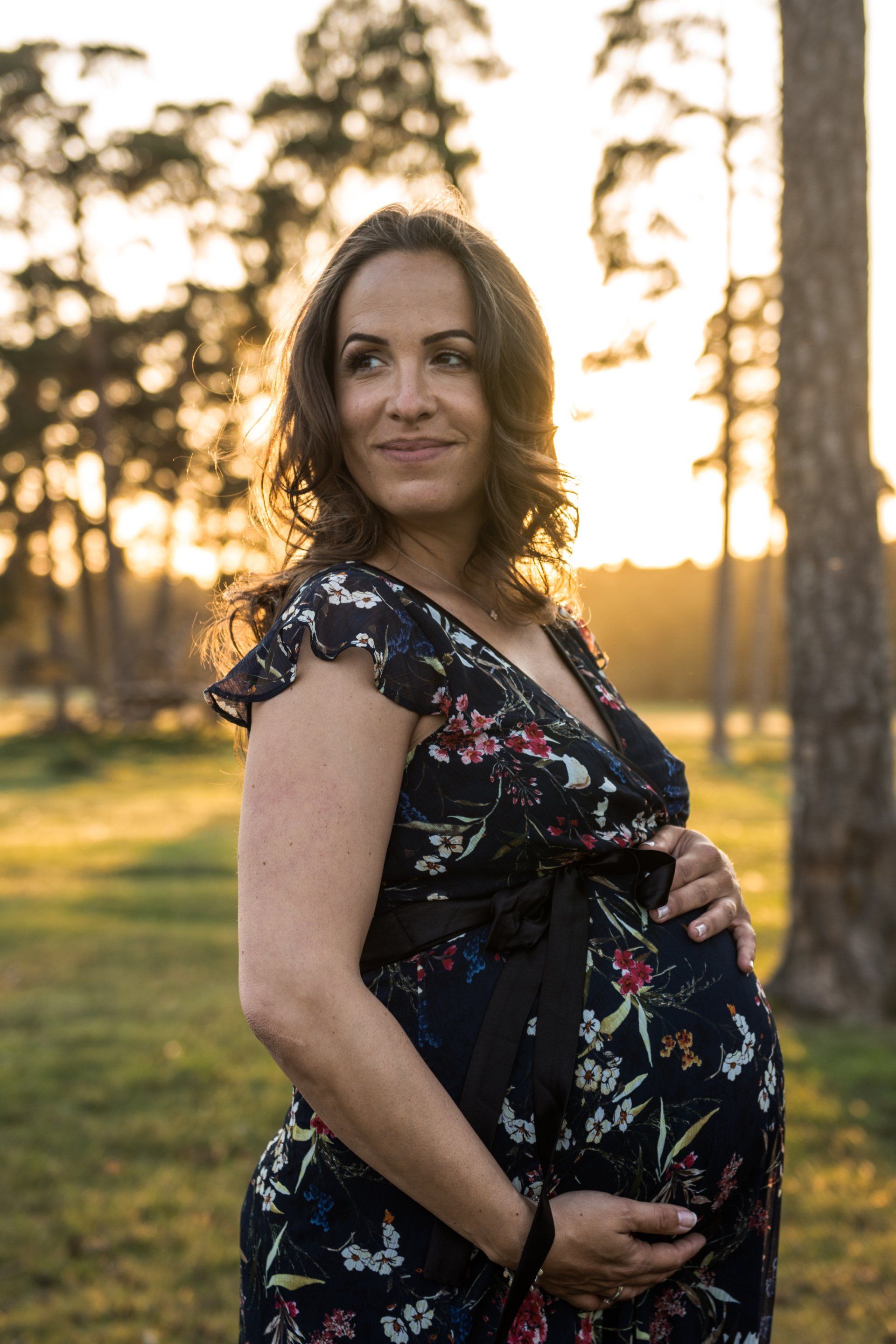 Schwangere Frau im geblümten Kleid, lächelt, hält Bauch, steht in einem sonnenbeschienenen Wald.