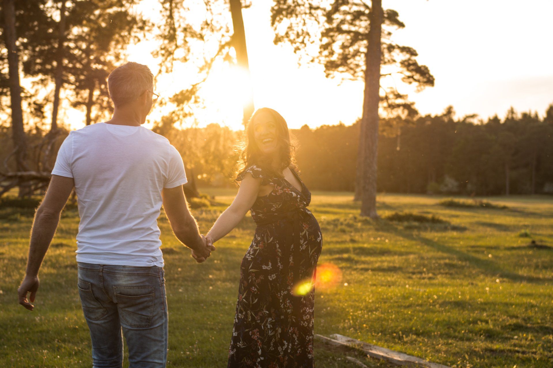 Schwangere Frau und Mann gehen Händchen haltend durch ein sonnenbeschienenes Feld und lächeln.