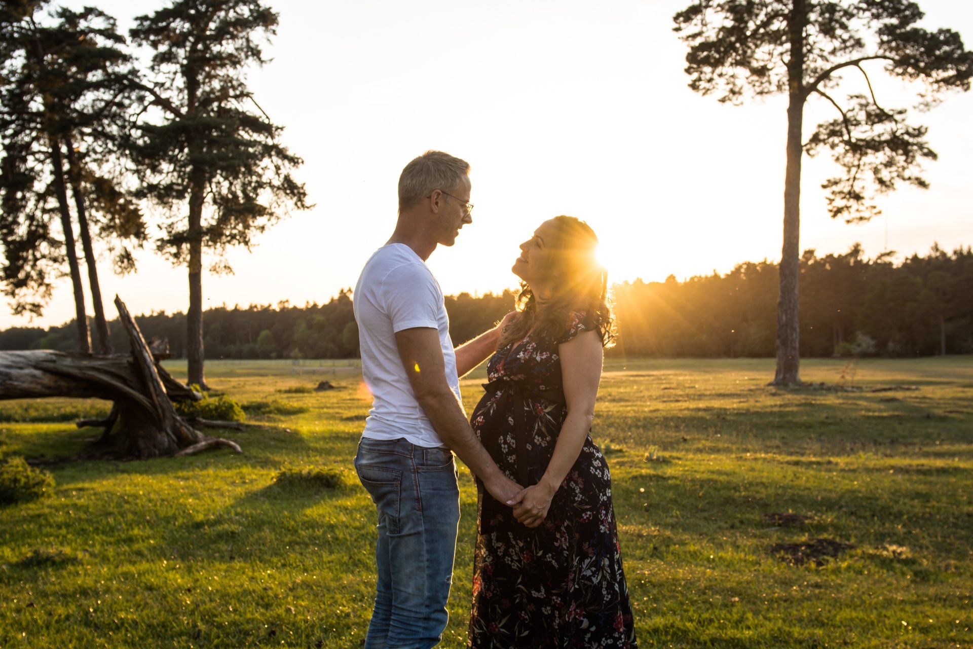 Schwangere Frau und Partner halten Händchen, im Gegenlicht des Sonnenuntergangs auf einem Feld.