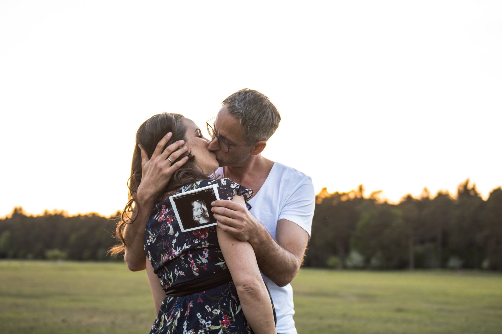Paar küsst sich, hält ein Ultraschallbild, feiert Schwangerschaft auf einem Feld bei Sonnenuntergang.