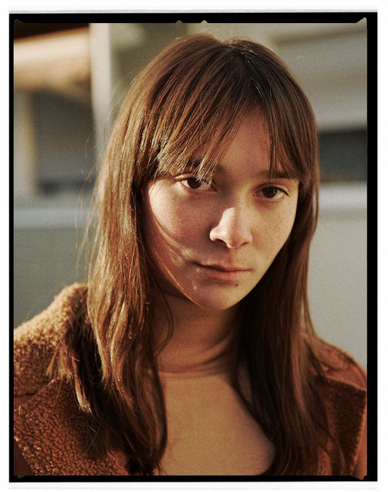 Frau mit langen braunen Haaren, Pony und ernstem Gesichtsausdruck, trägt einen braunen Mantel, im Freien.