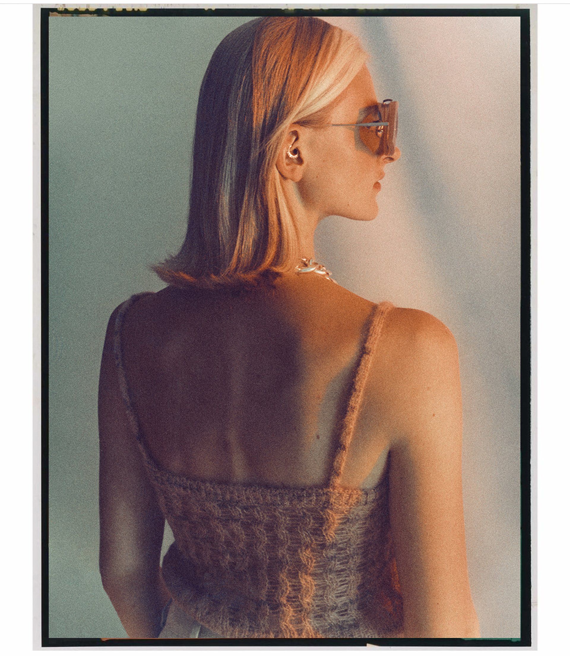 Frau mit Sonnenbrille, abgewandt, trägt ein strukturiertes Oberteil, kurzes blondes Haar, vor hellem Hintergrund.