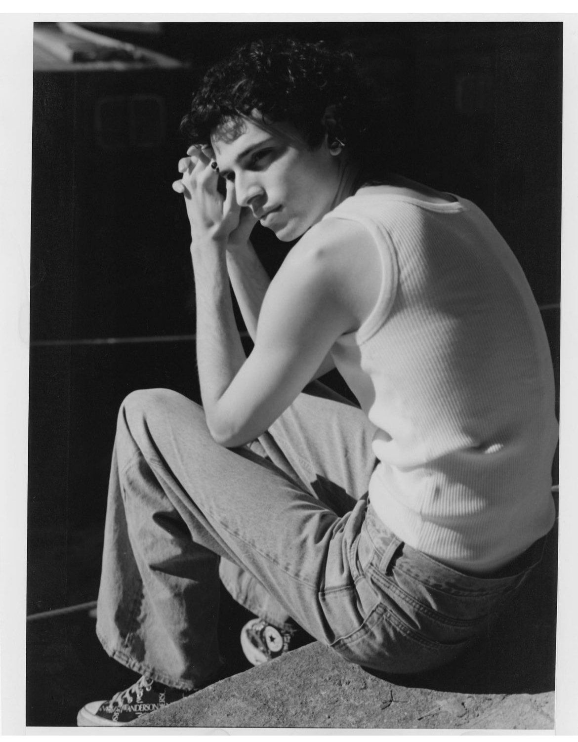 Mann sitzt mit verschränkten Armen und trägt ein weißes Tanktop, Jeans und Converse-Schuhe. Schwarzweißfoto.