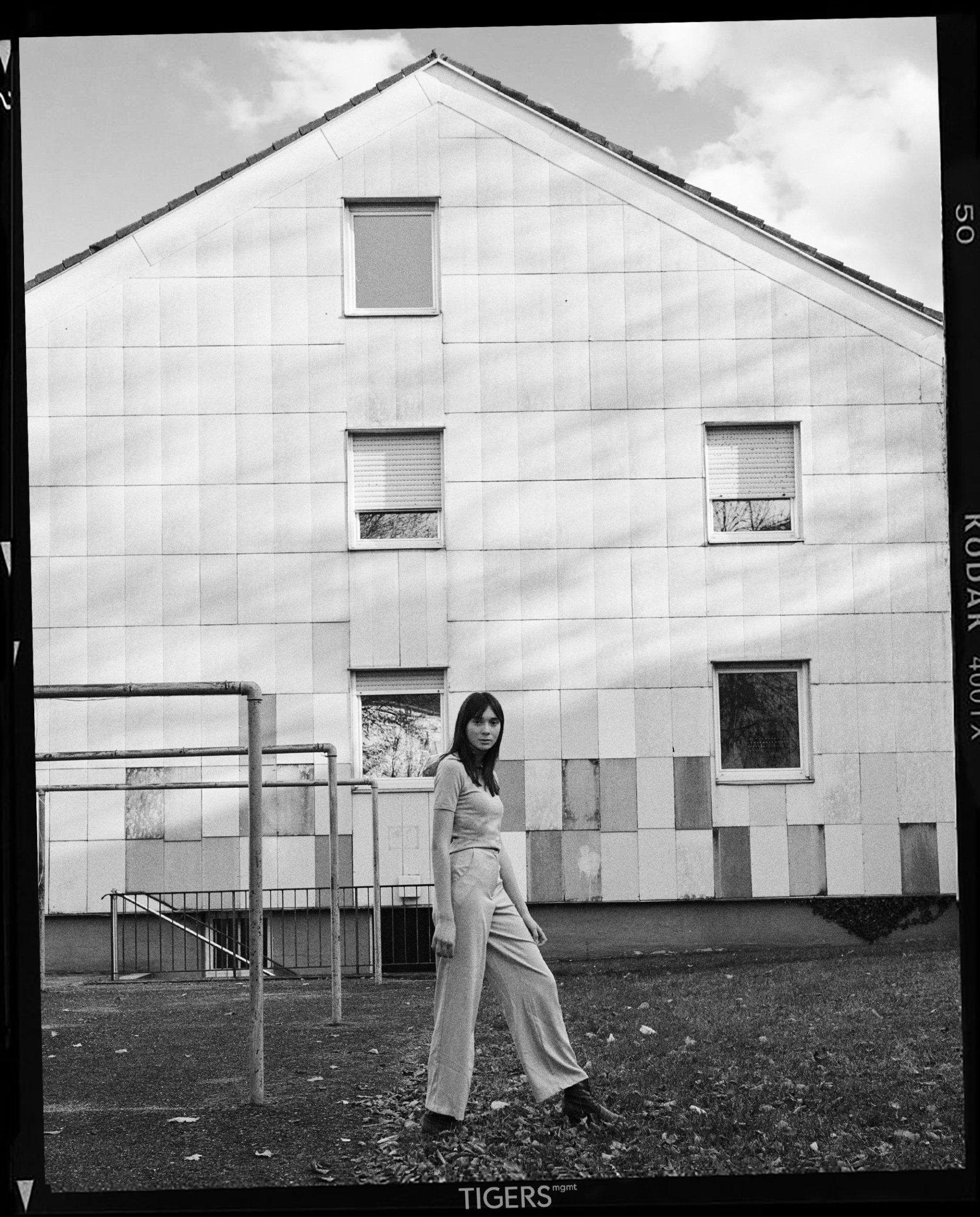 Frau im Overall steht vor einem verwitterten Gebäude mit Fenstern und einem kleinen Spielplatz.