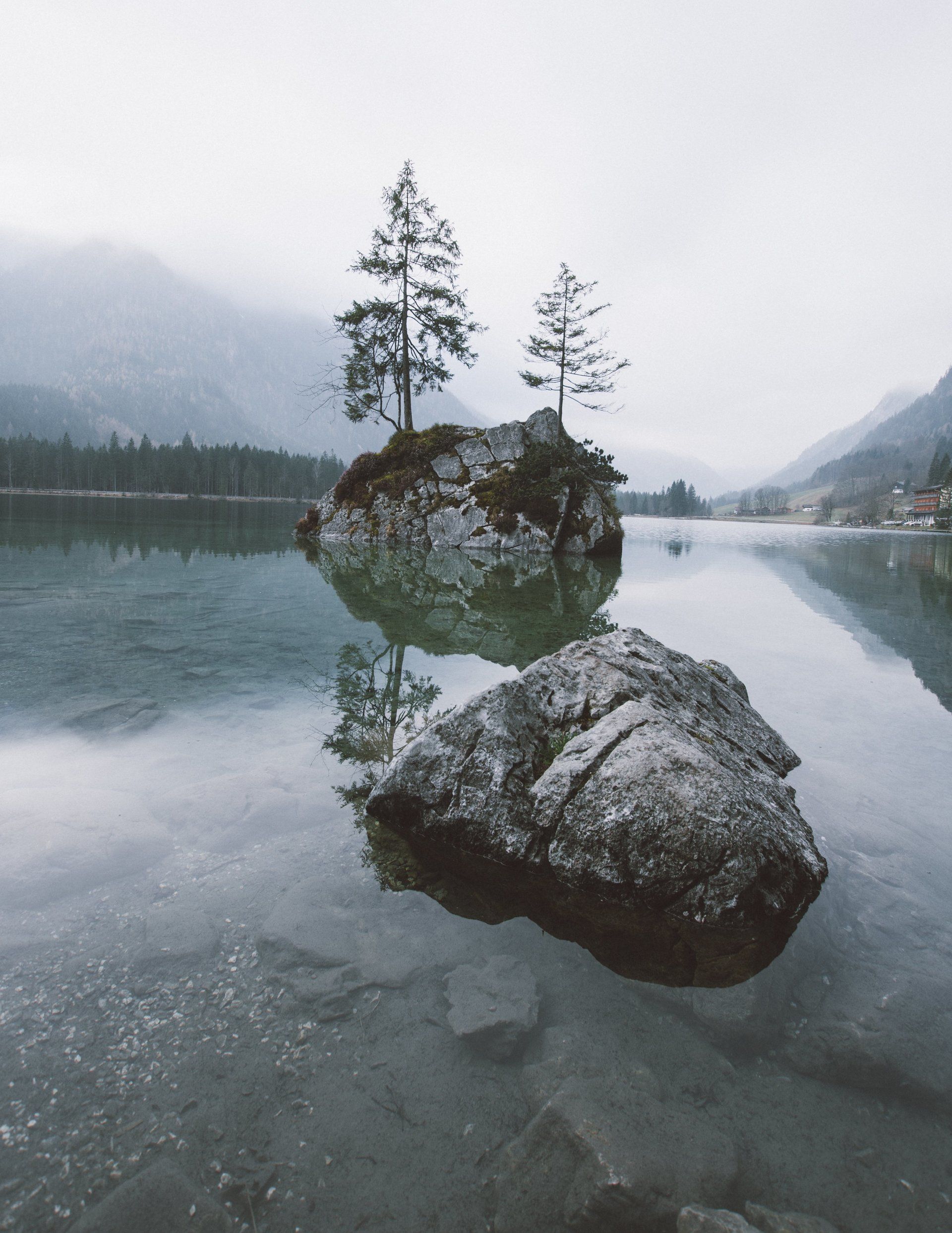 Felsige Insel mit zwei Bäumen, die sich im ruhigen See spiegeln. Grauer, bewölkter Himmel, Berge in der Ferne.