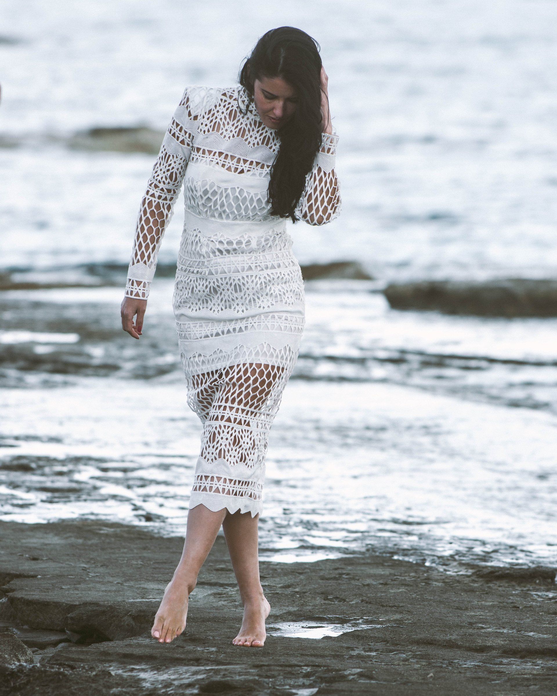 Frau im weißen Spitzenkleid steht barfuß auf Felsen in der Nähe des Ozeans.
