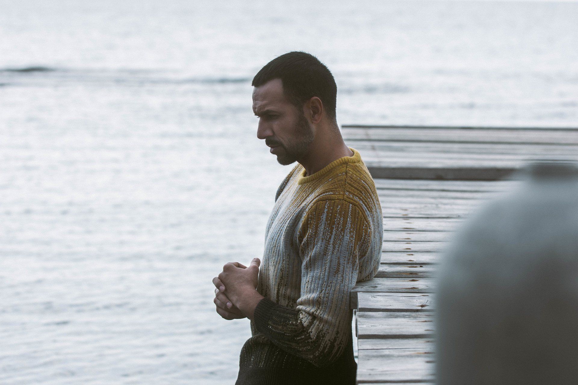 Mann auf einem Holzsteg, nachdenklich nach unten blickend, die Hände gefaltet. Grau-gelber Pullover, trübes Wasser.