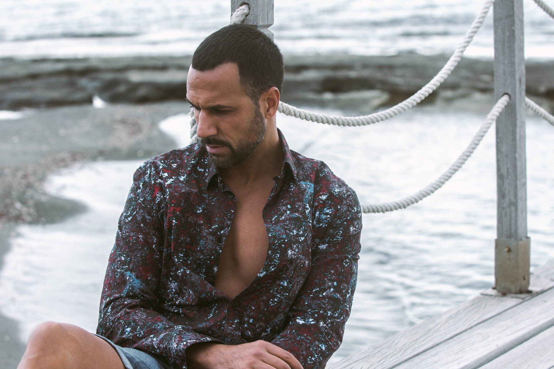 Mann sitzt auf einem Pier und blickt nach unten. Er trägt ein gemustertes Hemd und Shorts, im Hintergrund ist der Strand.