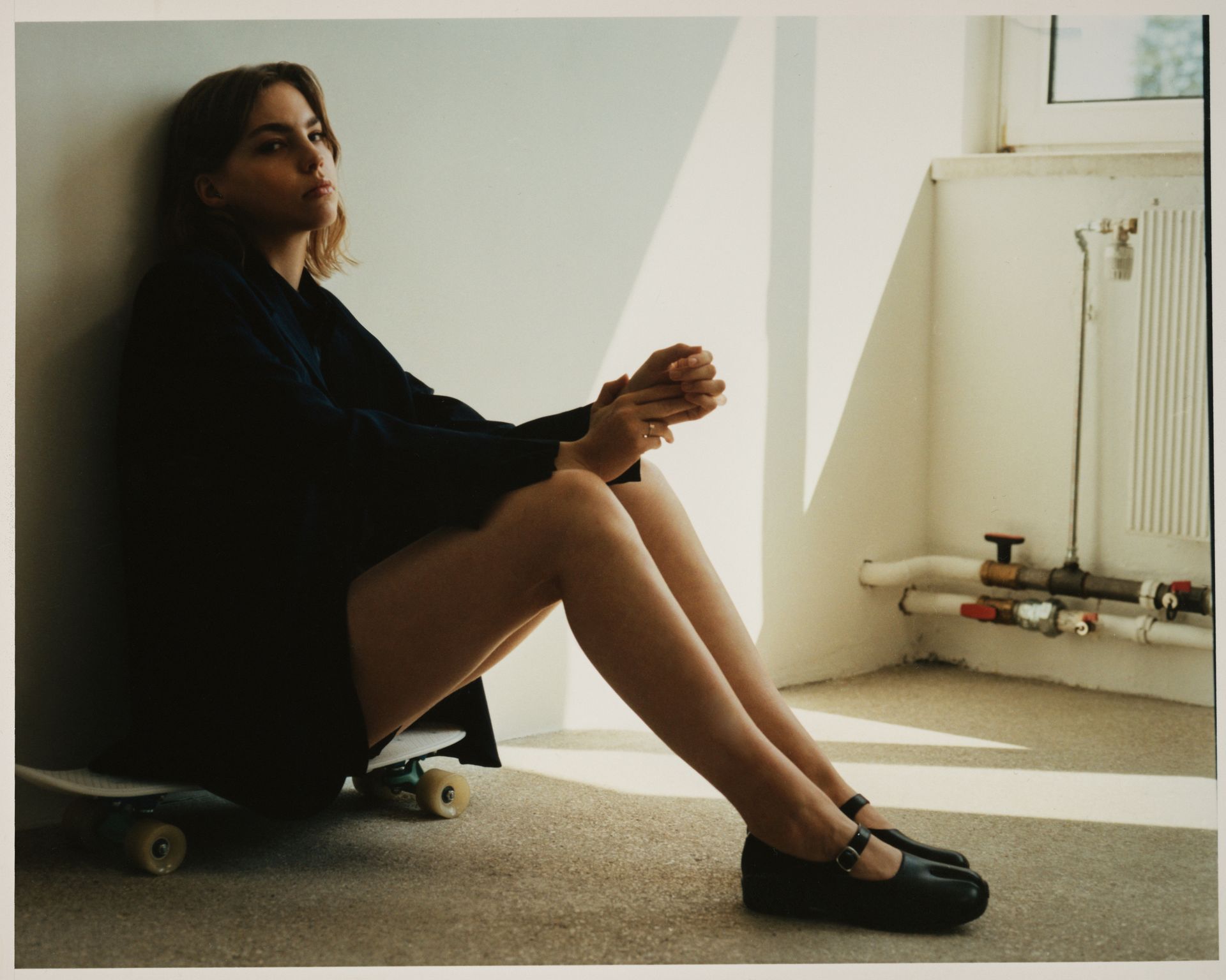 Frau sitzt mit gefalteten Händen an einer Wand. Sie trägt ein dunkles Oberteil und Schuhe und sitzt auf einem Skateboard.