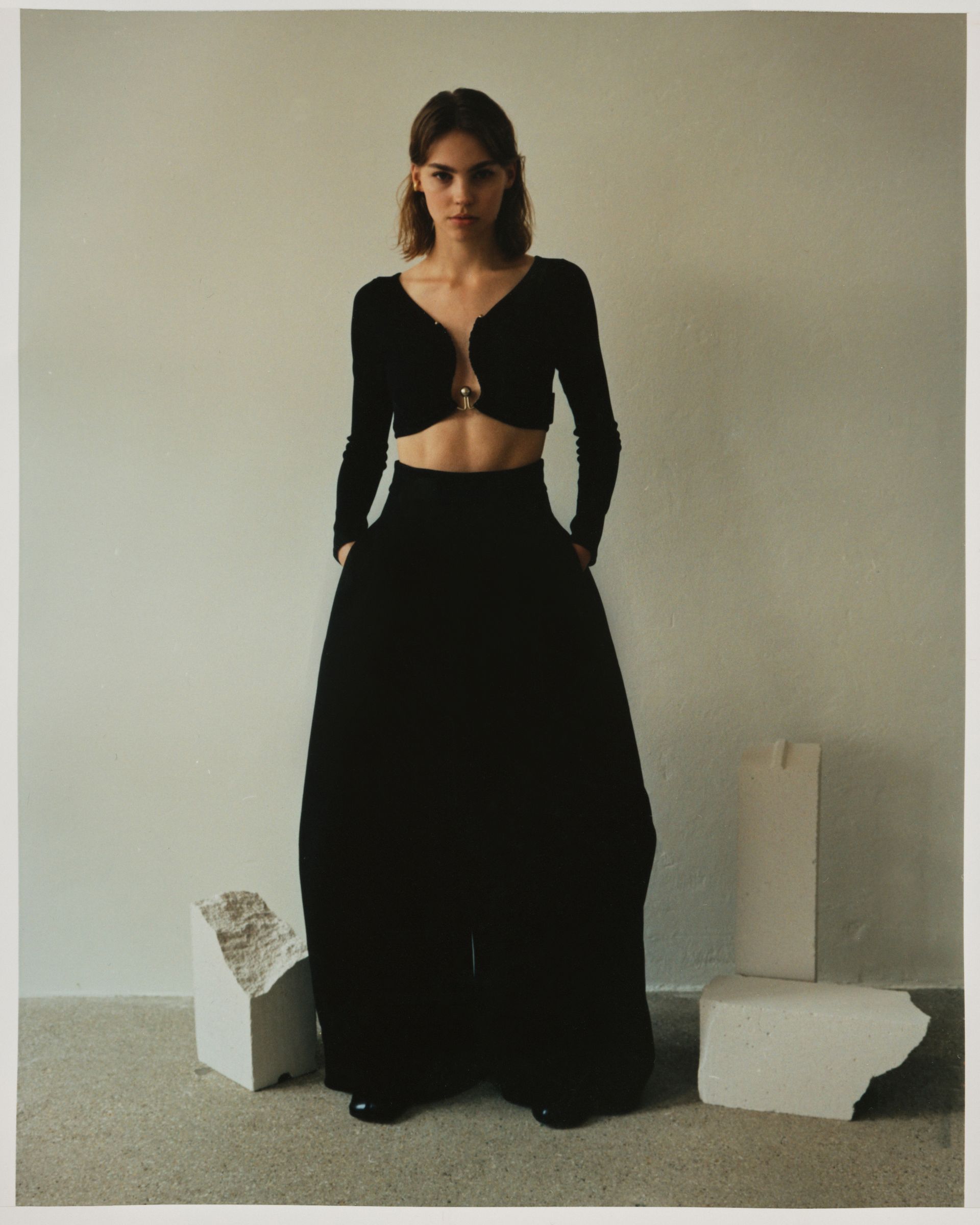 Frau in schwarzem bauchfreiem Top und weitem Rock steht im Studio mit abstrakten Steinblöcken.