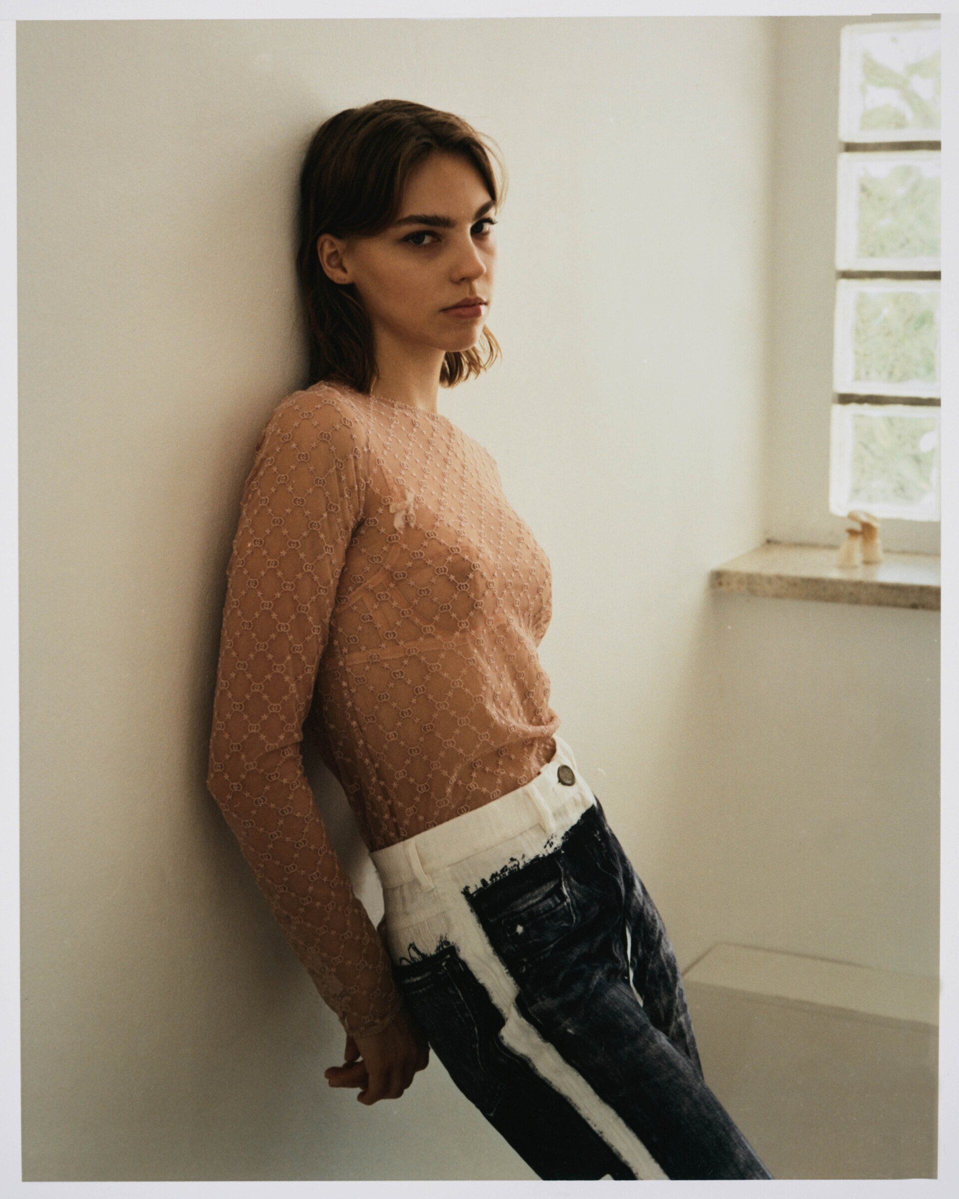 Frau in durchsichtigem Top lehnt an einer Wand, trägt Jeans und blickt in die Kamera neben einem Fenster.