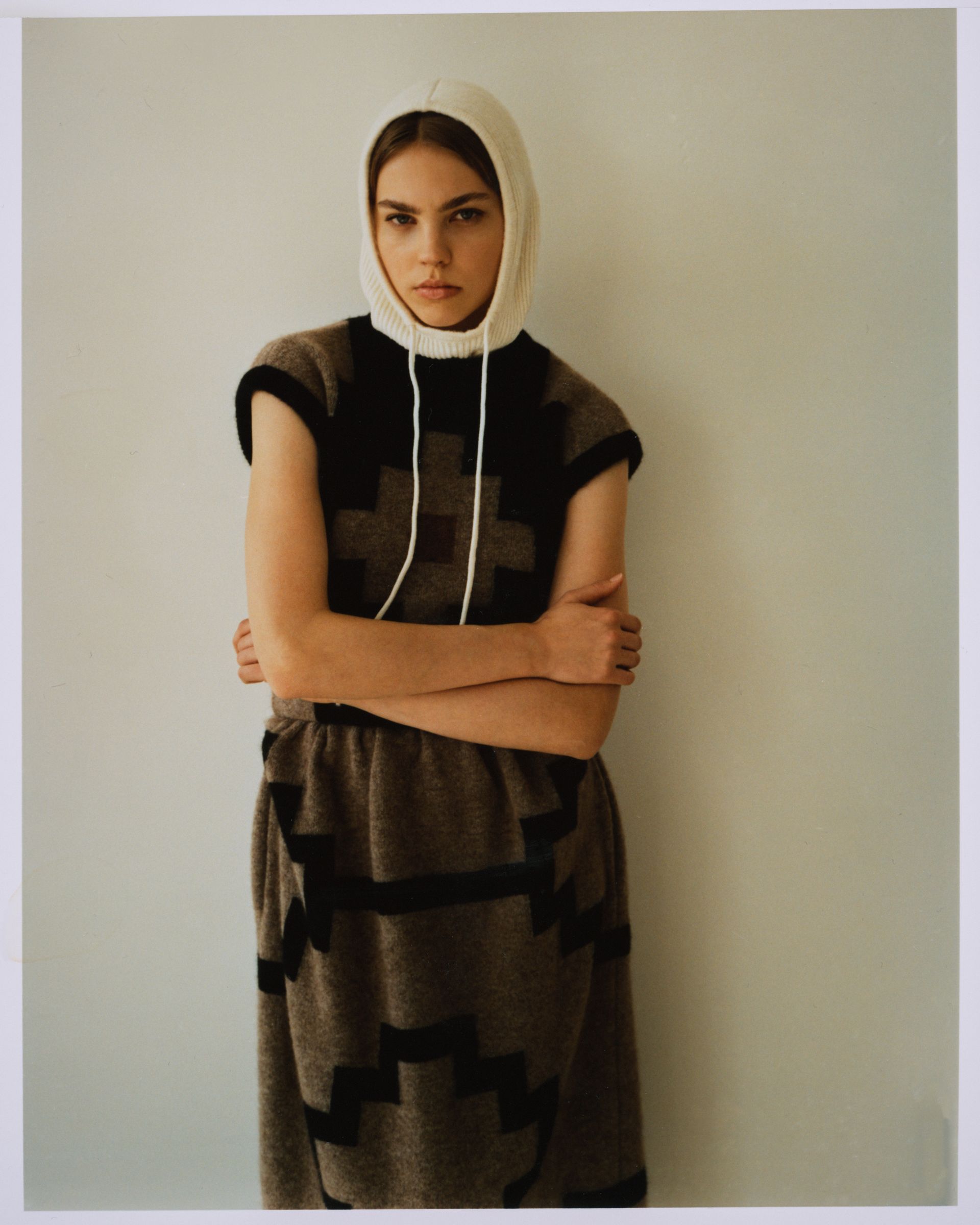 Frau in gemustertem Strickkleid mit Kapuze und verschränkten Armen steht vor einer schlichten Wand.