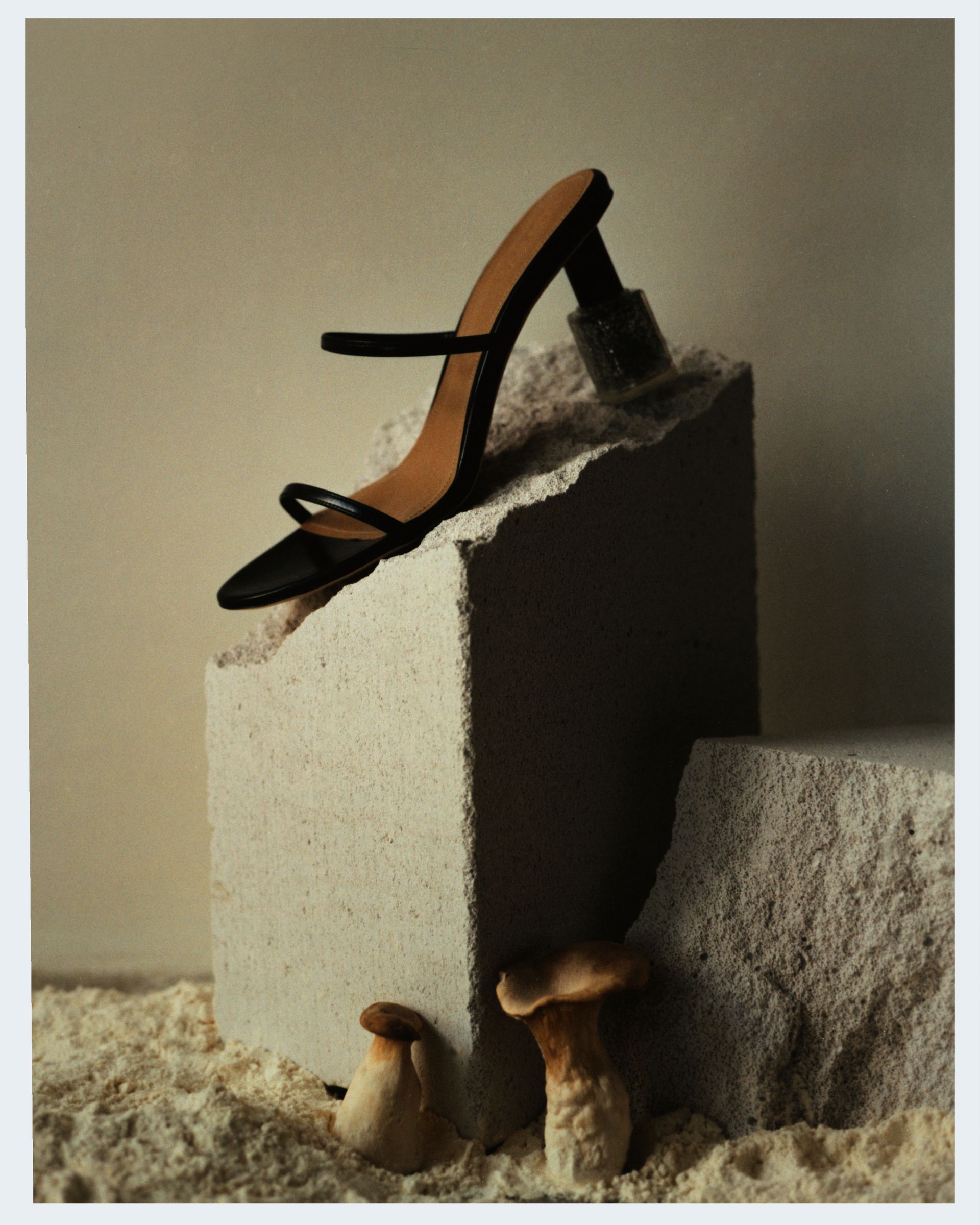 Schwarze Sandale mit Absatz auf einem strukturierten Block mit Pilzen, sandigem Untergrund und neutralem Hintergrund.