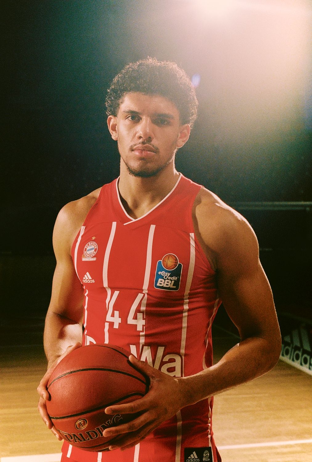 Basketballspieler in roter Uniform, hält Ball, auf einem Platz, blickt in die Kamera.