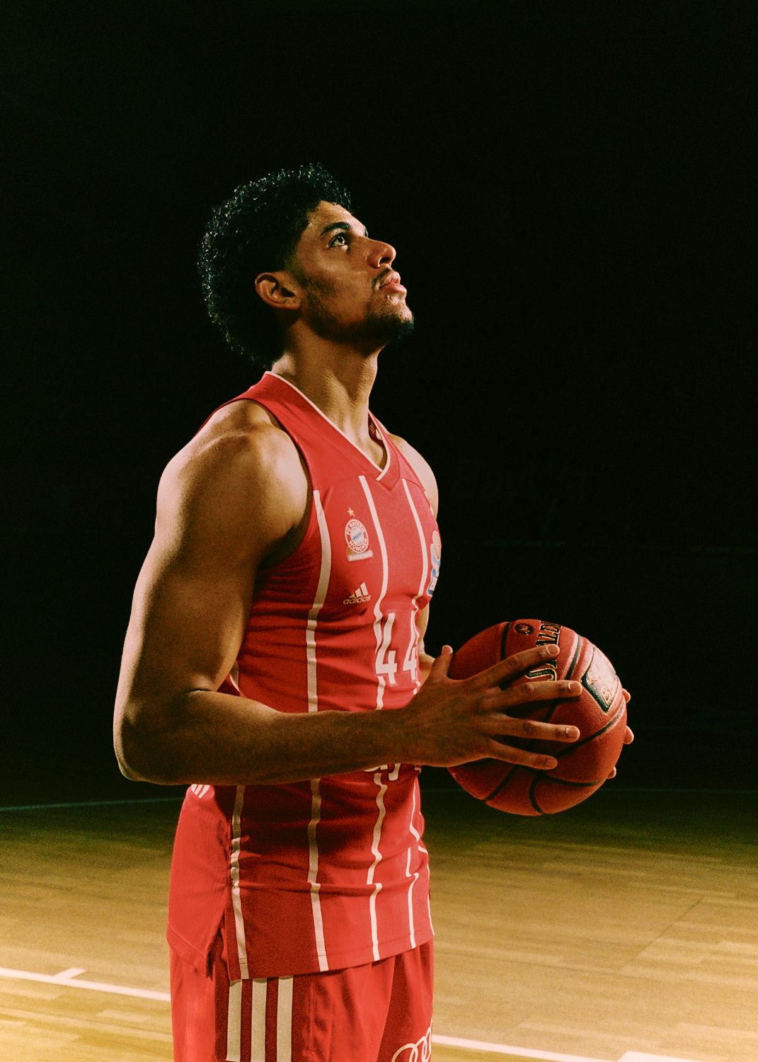 Basketballspieler in roter Uniform blickt nach oben, hält Ball, beleuchtet vor dunklem Hintergrund.