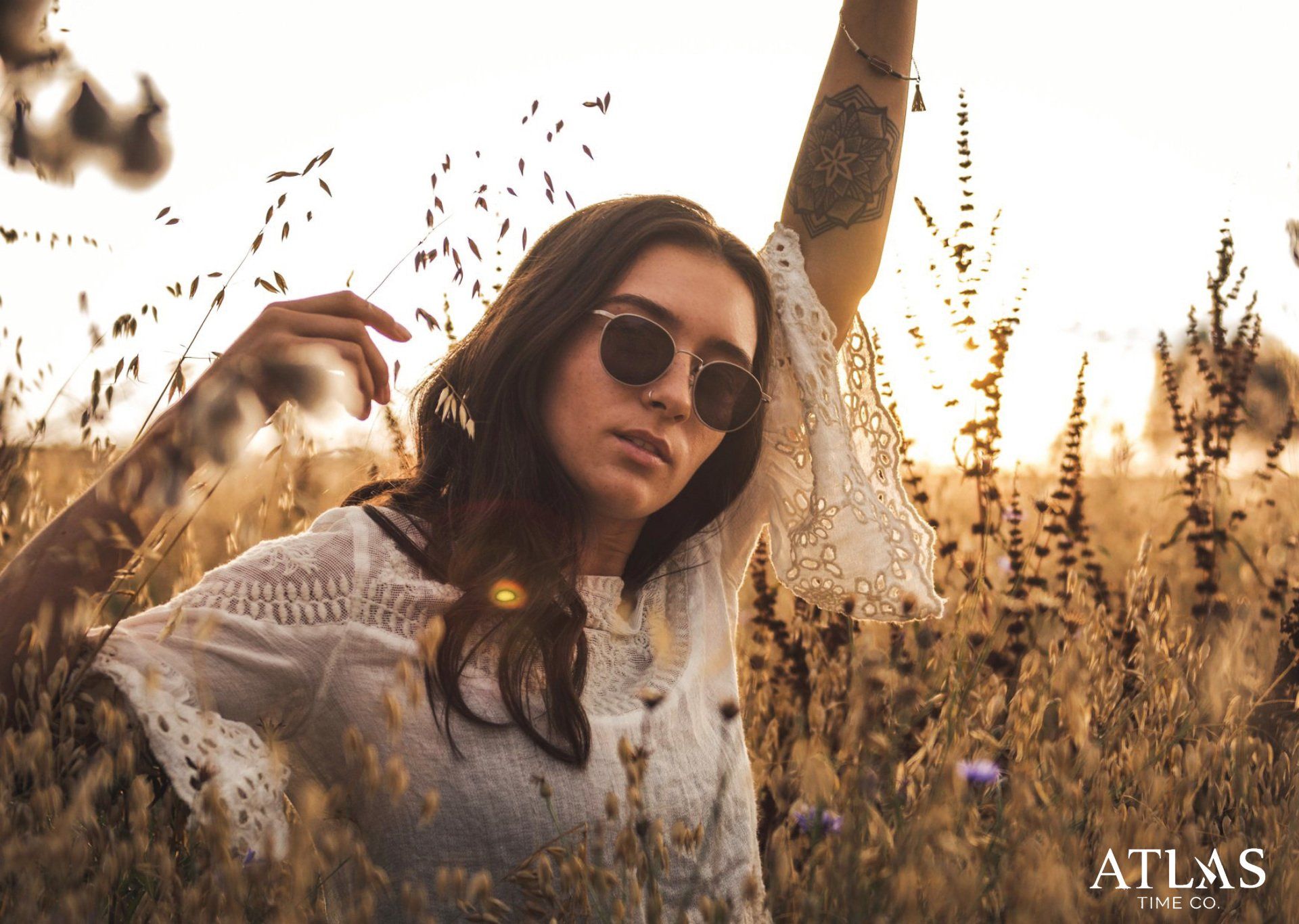 Frau mit Sonnenbrille und weißem Spitzentop im hohen Gras bei Sonnenuntergang.