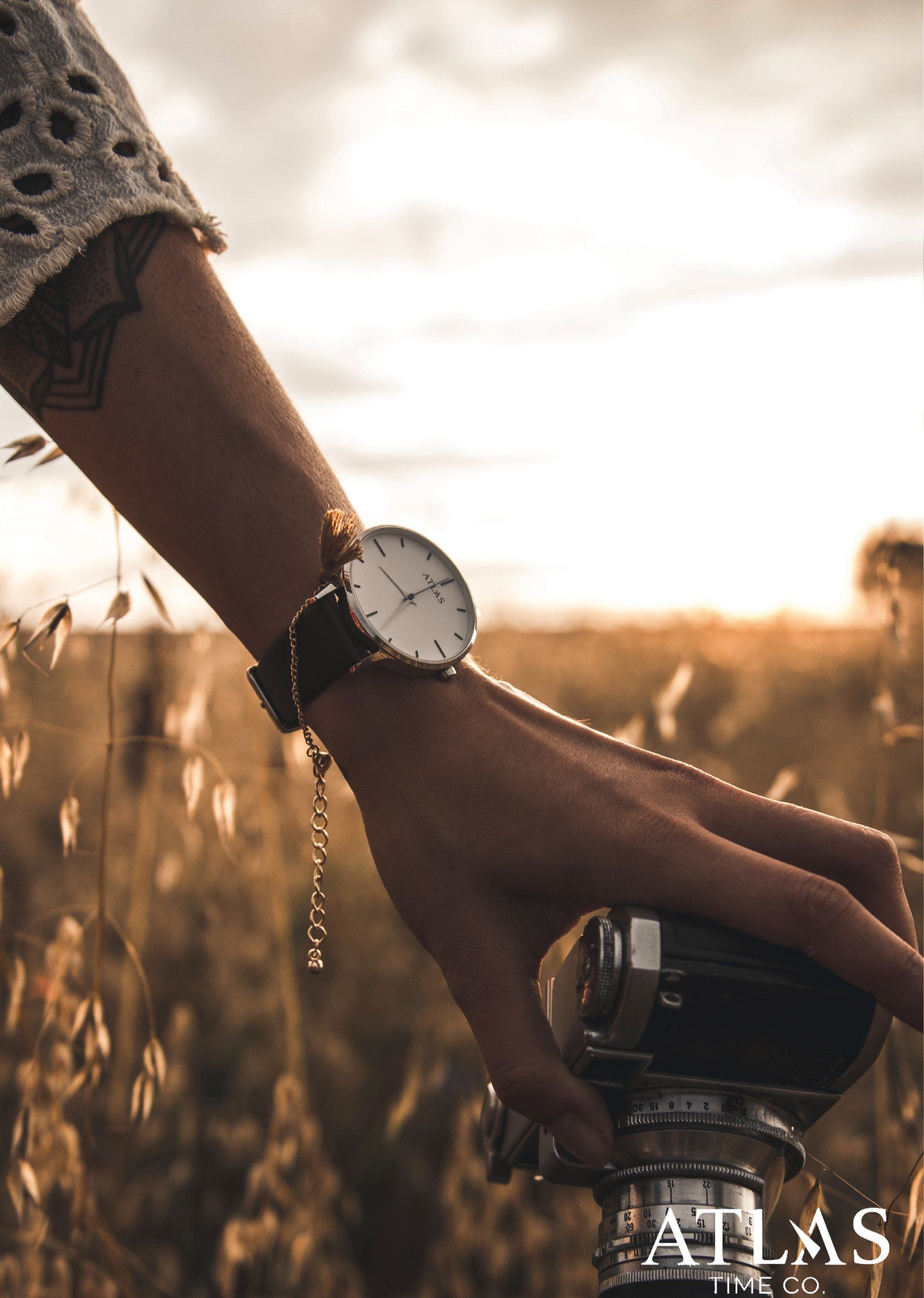 Arm einer Person mit Uhr, die eine Vintage-Kamera bei Sonnenuntergang auf einem Feld hält.