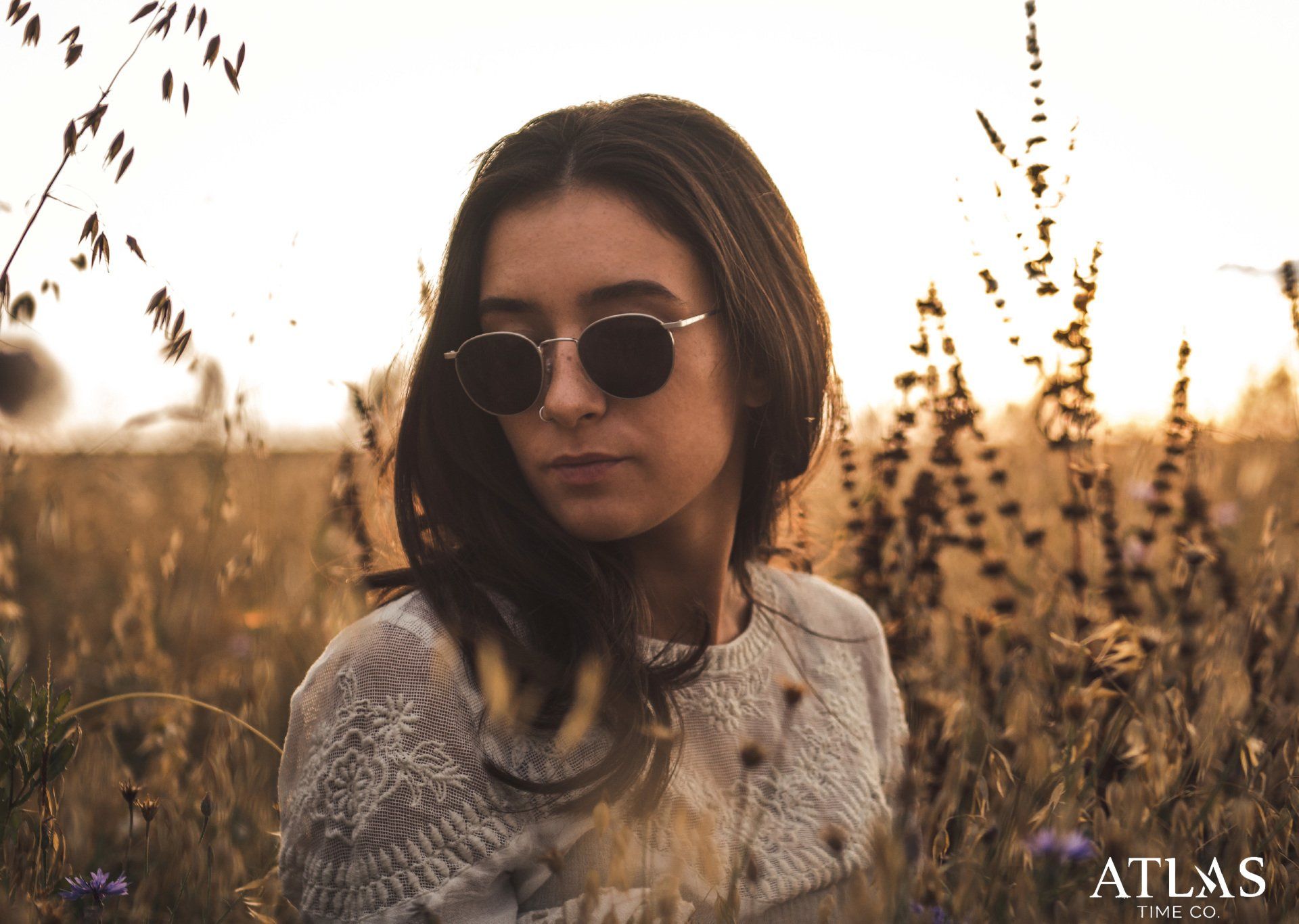 Frau mit Sonnenbrille steht im hohen Gras, beleuchtet von goldenem Sonnenlicht.