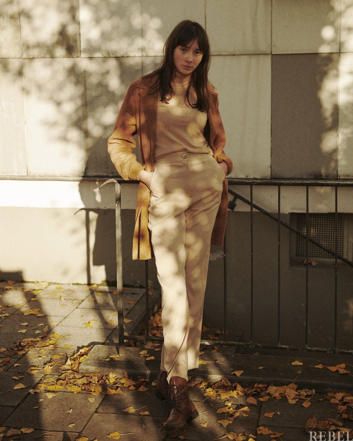 Frau in hellbraunem Outfit posiert im Freien, Hände in den Taschen, im Herbstschatten.