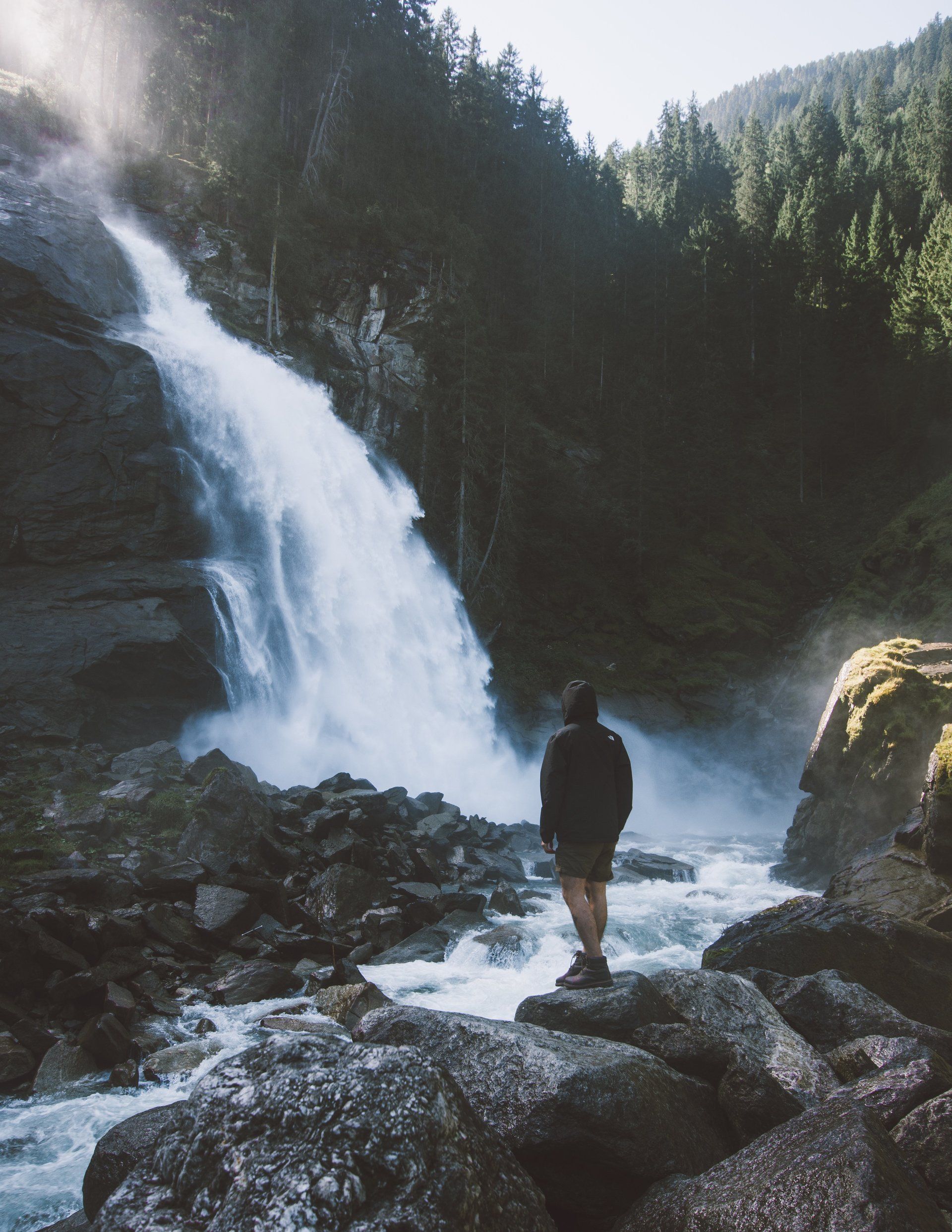 Person steht vor einem Wasserfall in einem üppigen grünen Wald. Sonnenlicht scheint und erzeugt Nebel.