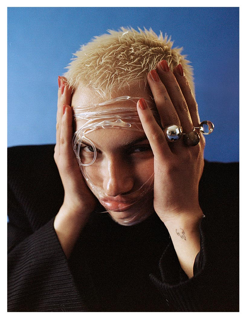 Person mit blondem Haar, in Plastikfolie gehüllt, Hände im Gesicht, rote Nägel, blauer Hintergrund.