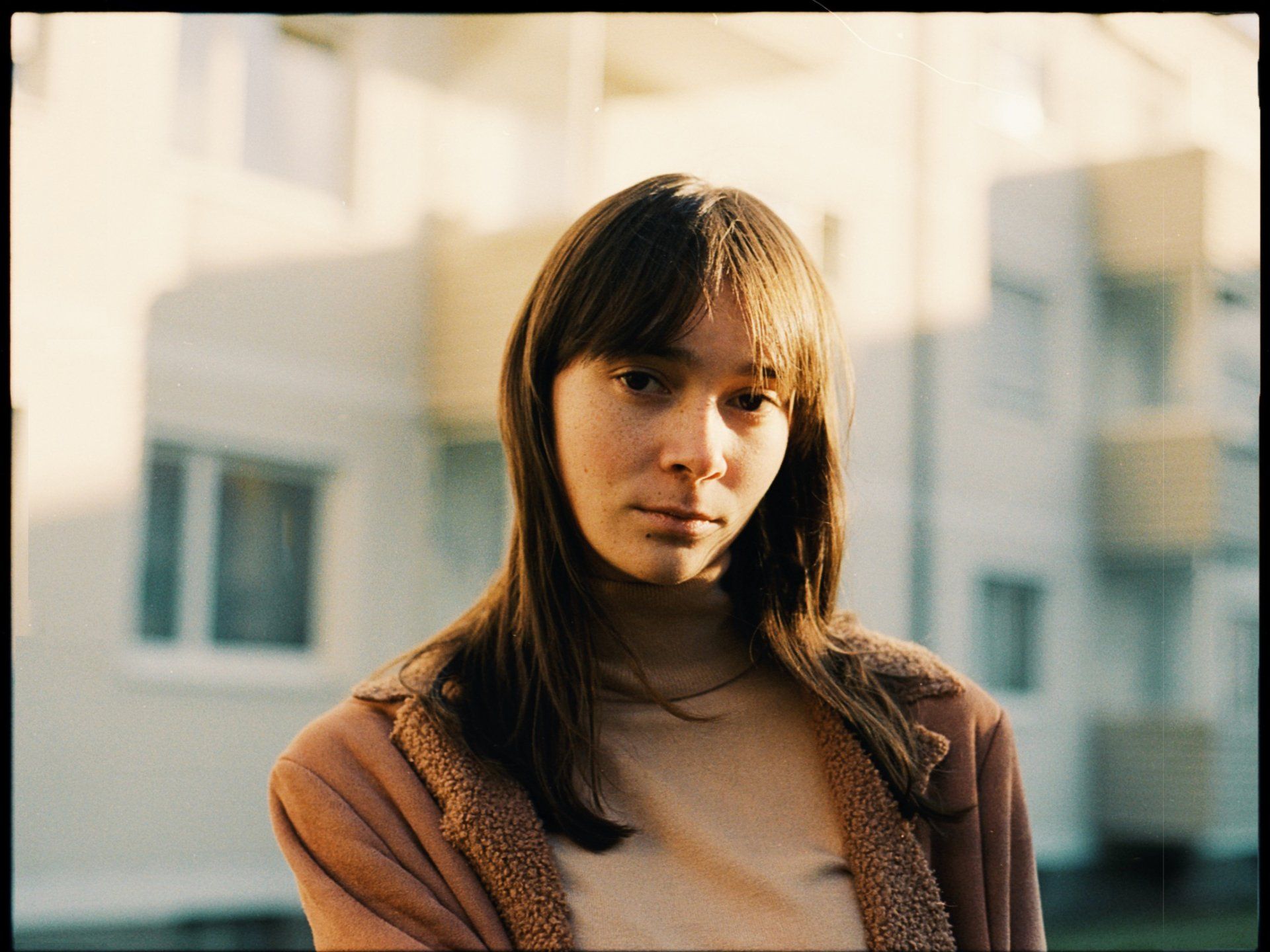 Frau mit langem braunem Haar und ernstem Gesichtsausdruck steht vor einem Gebäude und trägt eine beige Jacke.